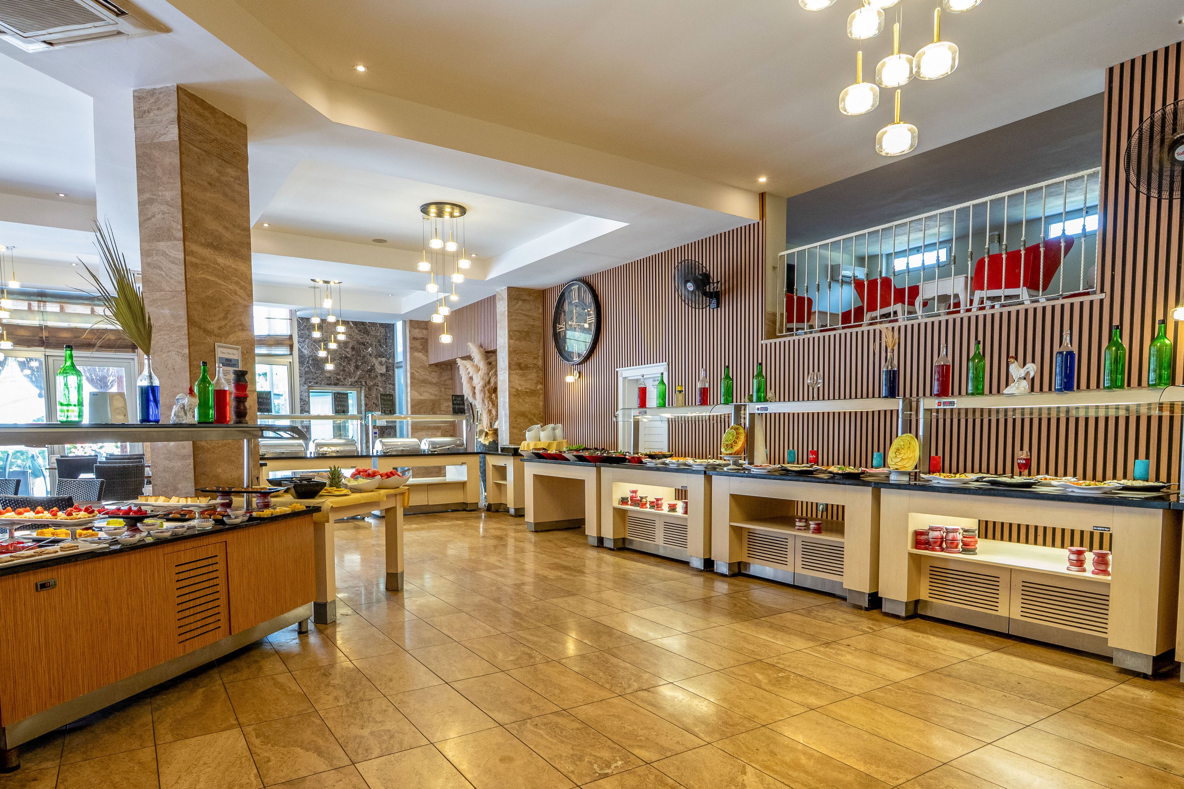 A spacious buffet restaurant interior features multiple counters arranged with plates, bowls, and colorful bottles. The setting appears to be a hotel dining area with tiled floors, warm lighting, and contemporary wooden finishes. Decorative glass bottles in various colors line the back counters, while a large wall clock and vertical slats add visual interest. Ceiling lights and natural light from windows create a bright, welcoming atmosphere suitable for self-service dining.