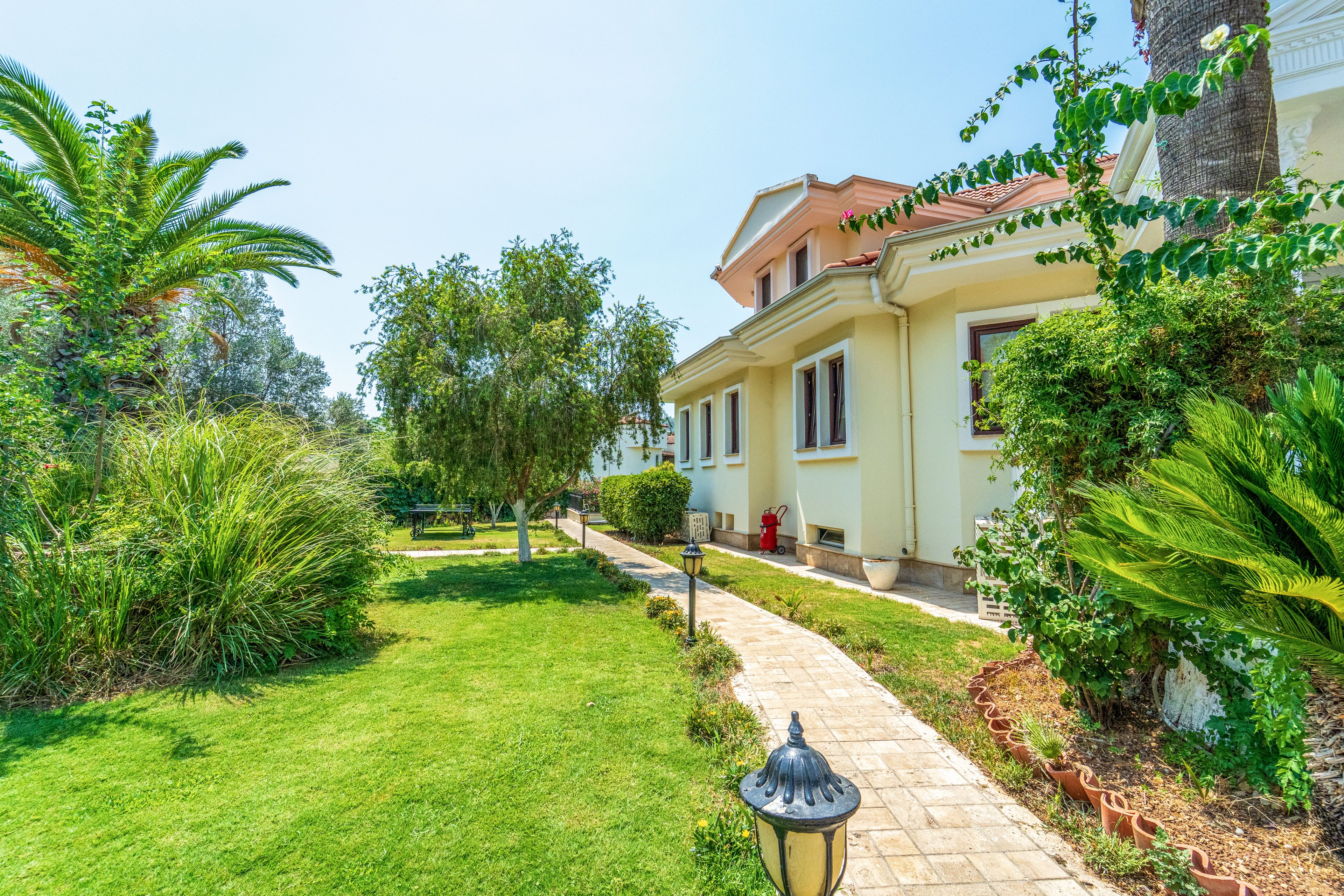 A bright, cream-colored villa is surrounded by a well-maintained, lush green garden. A stone pathway leads along the side of the house, bordered by trimmed grass, shrubs, and small trees. Tall palm trees and dense foliage create a Mediterranean feel under a clear blue sky. A decorative garden lamp and red fire hydrant add visual accents to the peaceful outdoor setting.