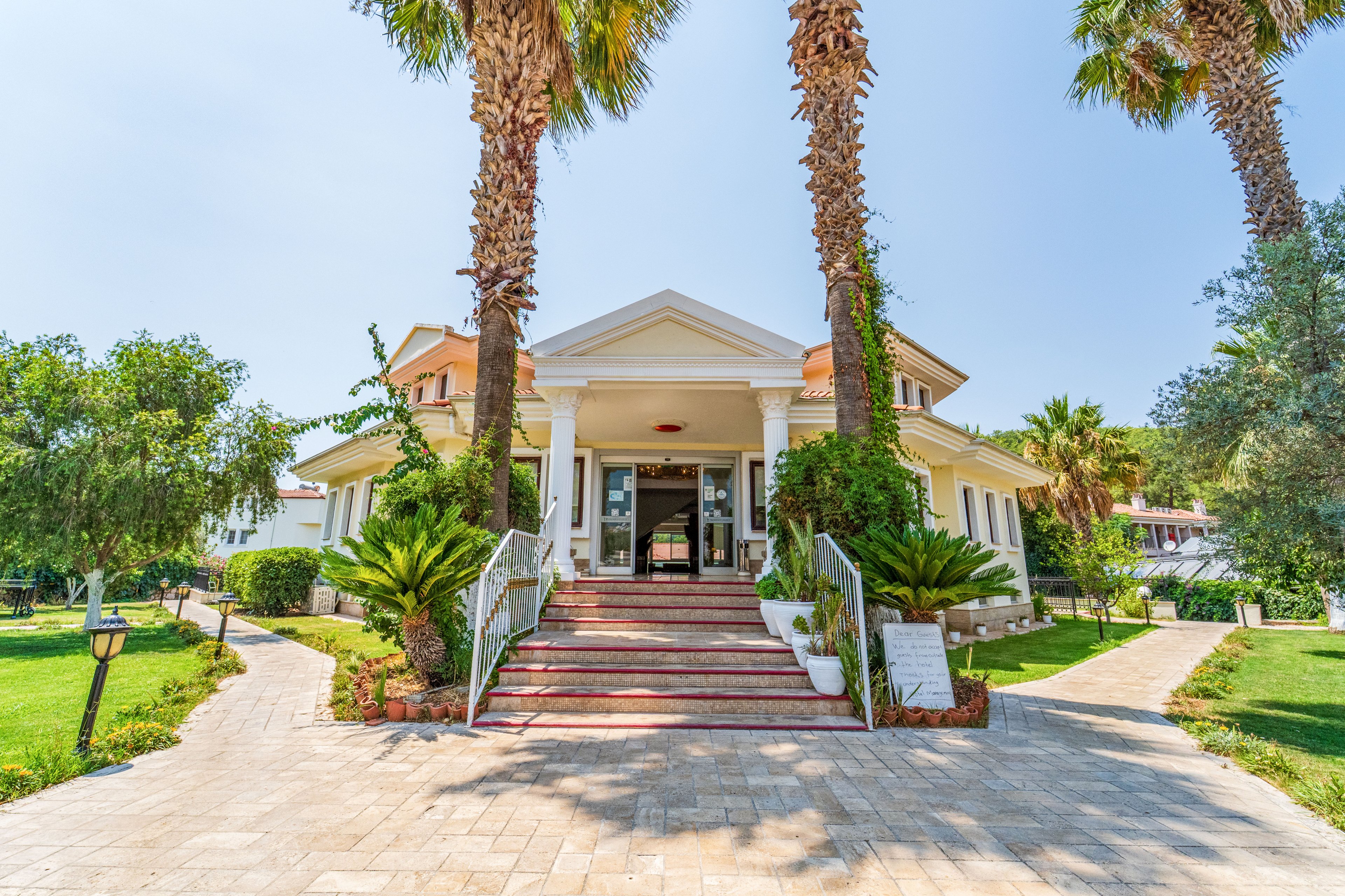 A bright Mediterranean-style villa stands at the end of a tiled pathway lined with lush greenery and tall palm trees. Wide steps lead up to the main entrance, framed by white columns and a triangular pediment. The surrounding garden features manicured shrubs, lawn areas, and outdoor lighting, suggesting a welcoming resort or guesthouse setting. Clear blue sky and strong sunlight create a warm, inviting atmosphere.
