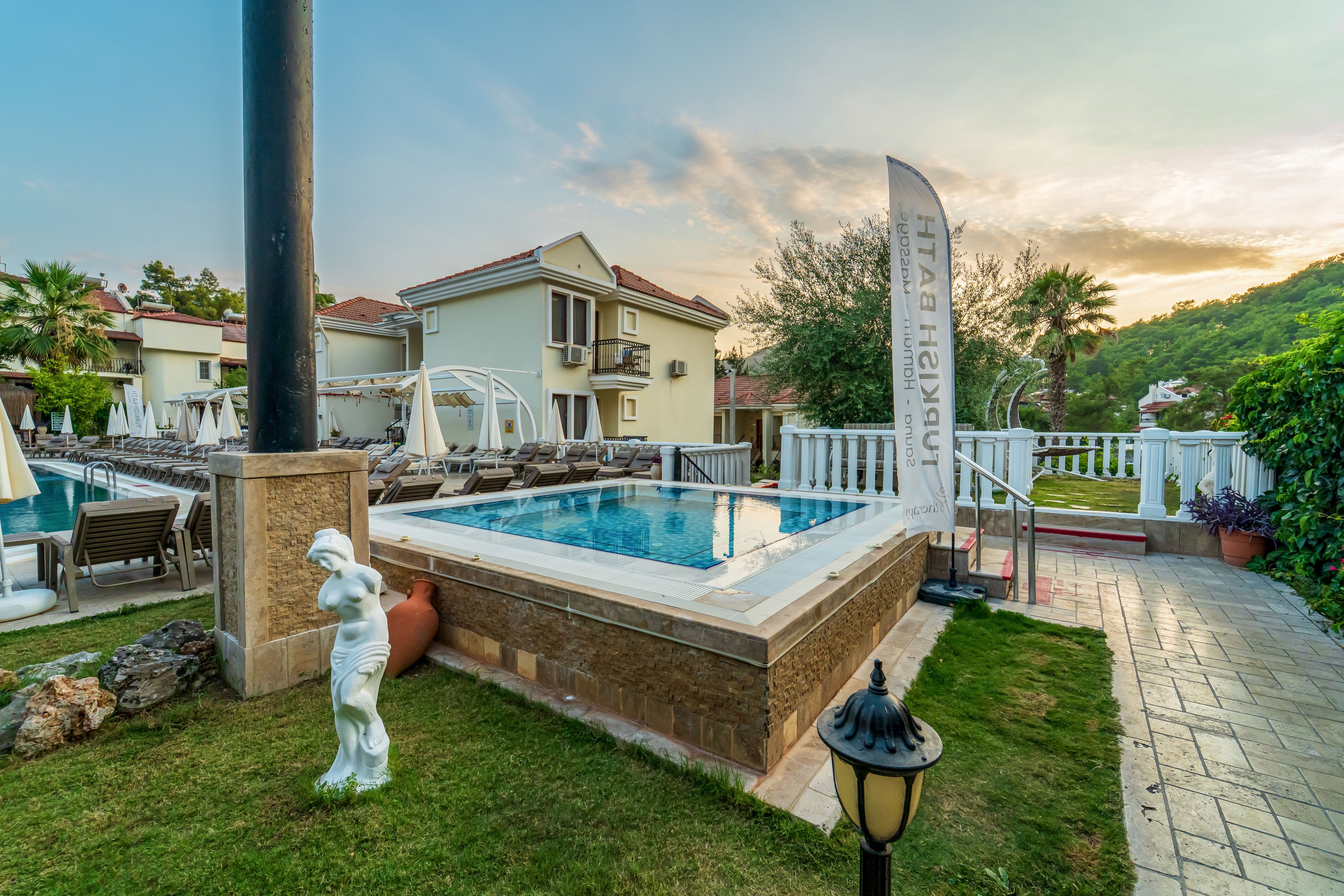 An elevated swimming pool sits beside a landscaped garden in front of a light-colored villa at a resort. White lounge chairs and umbrellas surround the pool area, with hills and greenery visible in the background. A decorative statue, pathway lamp, and stone details add elegance to the outdoor space. The sky is tinged with warm sunset colors, creating a relaxed vacation atmosphere.