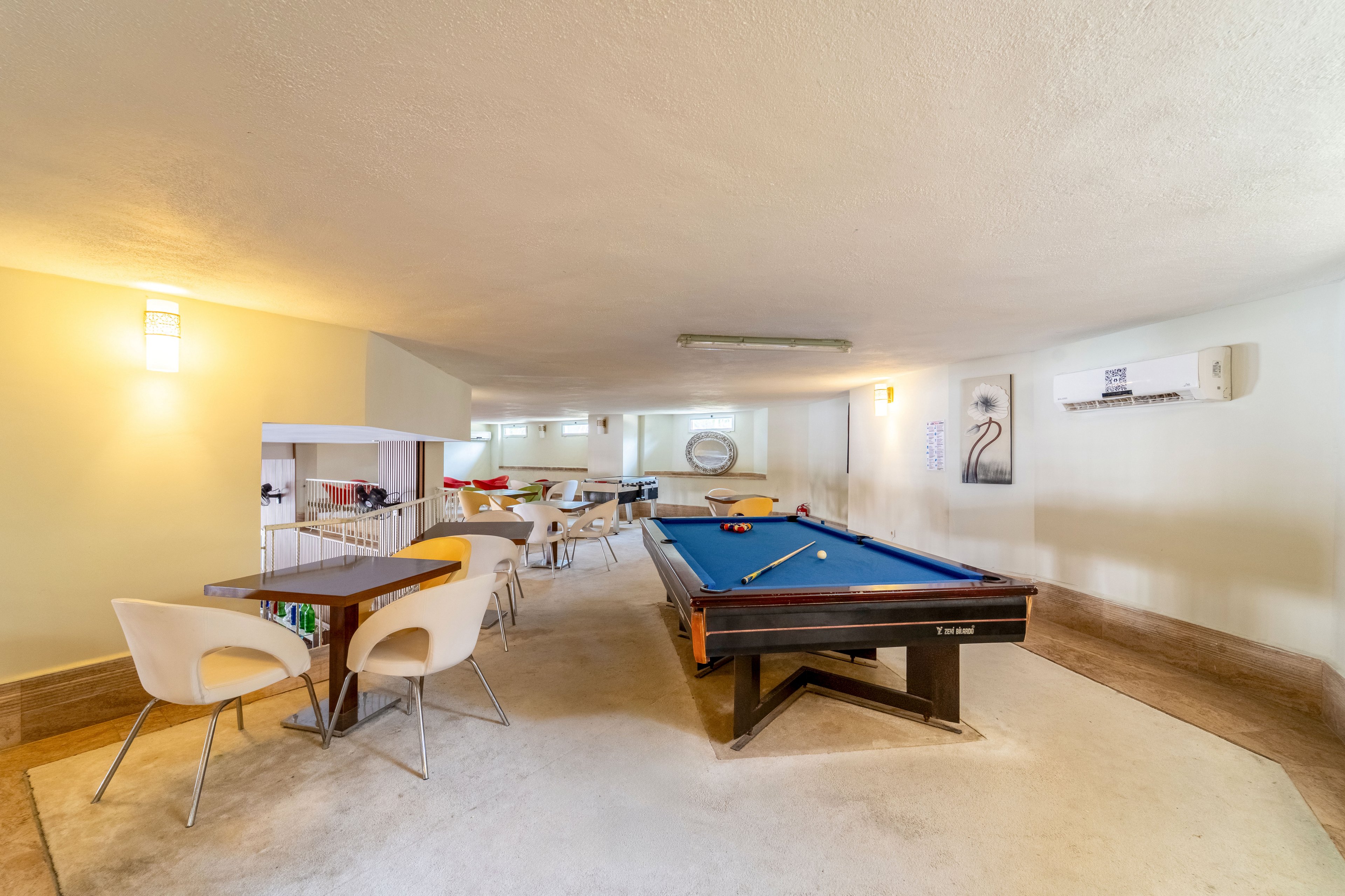 A spacious recreation room features a central blue-felt pool table with cues resting on top. Modern white chairs surround small tables, creating a casual seating area for socializing or games. The room is brightly lit with wall sconces and overhead lighting, and a large mirror reflects the interior, adding depth. Neutral walls and flooring give the space a clean, contemporary feel suitable for leisure and relaxation.