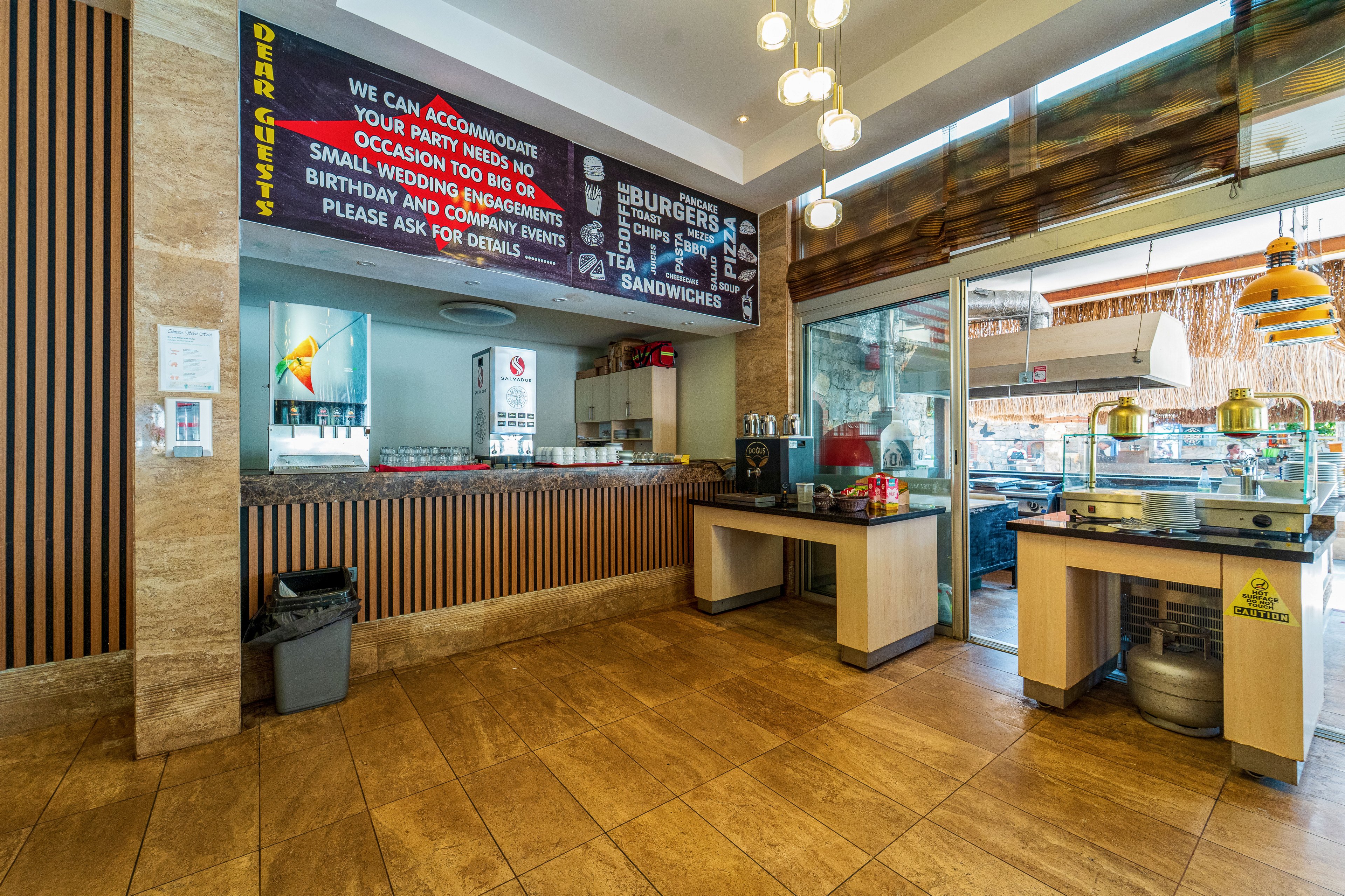 Wide-angle view of a casual burger restaurant interior featuring a service counter and food preparation area. A large black menu board above the counter lists items such as burgers, fries, and sandwiches, along with event accommodation information. The space includes tiled flooring, wood panel accents, hanging pendant lights, and a glass door leading to an outdoor area. Stainless steel kitchen equipment and display units are visible, suggesting a fast-casual dining environment.