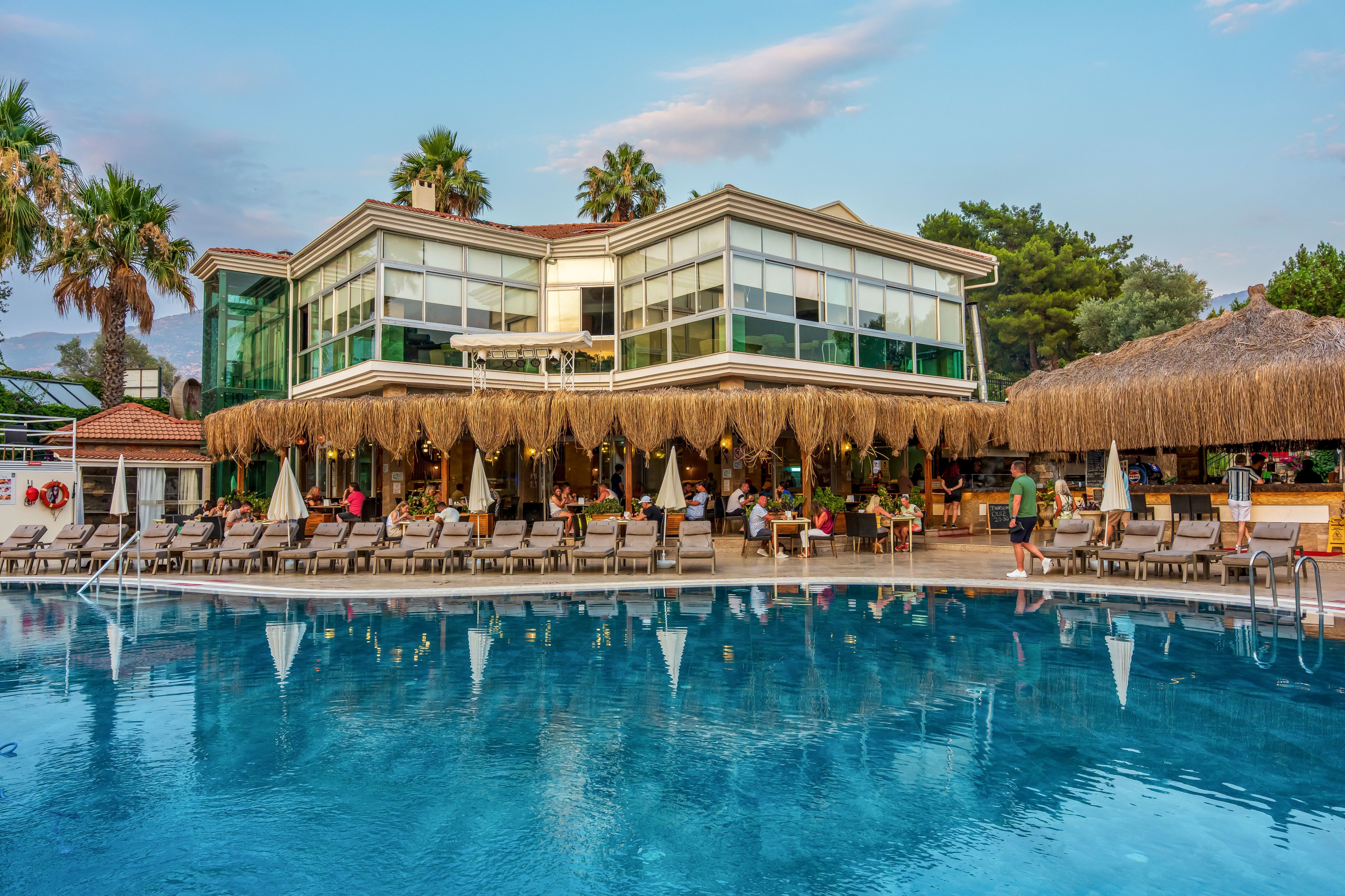 A modern resort hotel overlooks a large outdoor swimming pool in the early evening light. Guests relax on sun loungers and sit under a thatched-roof terrace beside the water. Tall palm trees and lush greenery frame the building, adding a tropical atmosphere. The calm blue pool surface reflects the hotel’s glass facade and the soft colors of the sky.