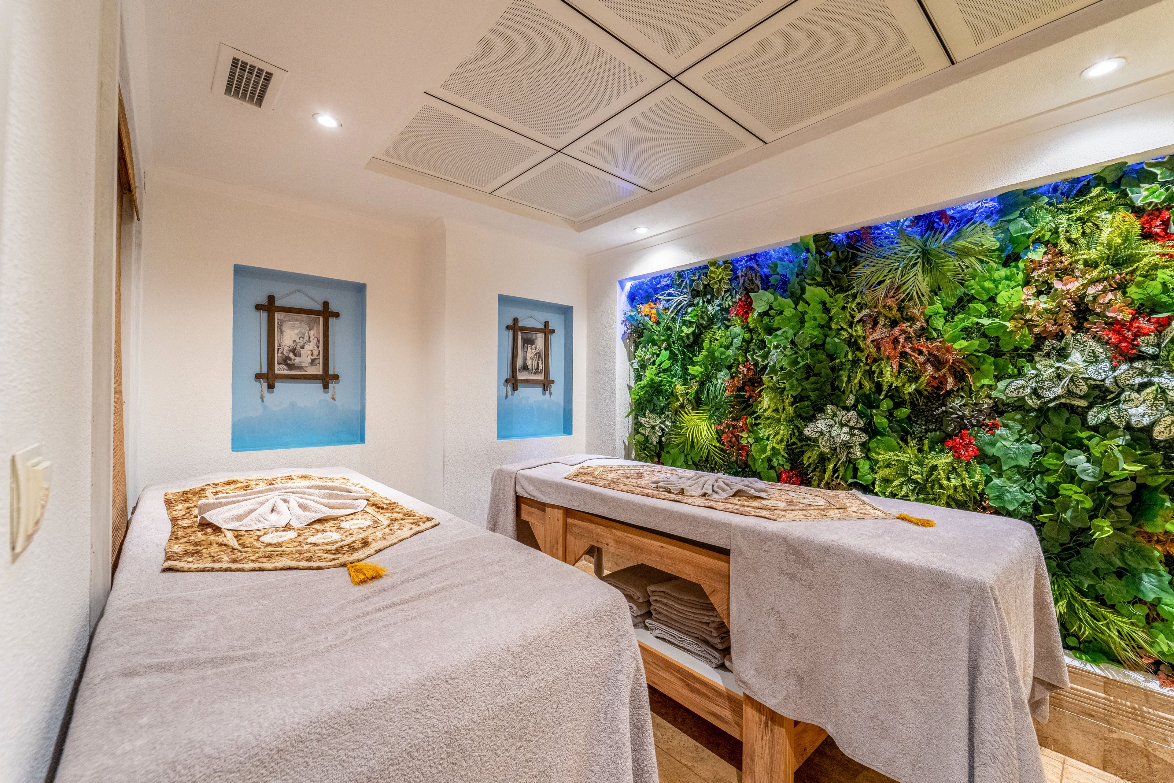 A cozy spa treatment room features two massage tables covered with soft gray linens and decorative towels. One wall is filled with a vibrant vertical garden of lush green plants and red flowers, adding a natural focal point. The space is brightly lit with recessed ceiling panels and has minimalist white walls accented by framed artwork. The overall atmosphere feels calm, clean, and inviting, ideal for relaxation and wellness treatments.