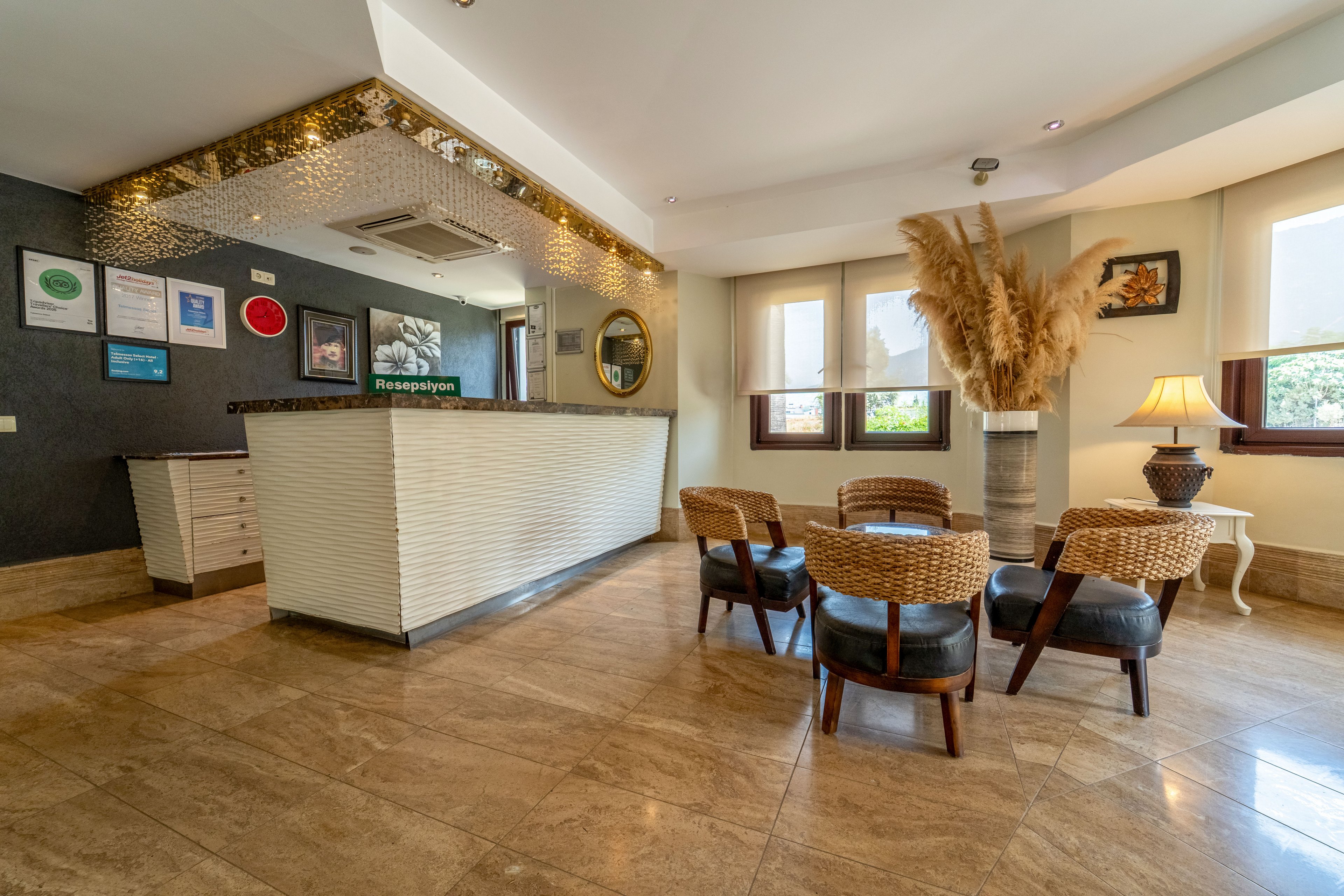 A contemporary hotel reception desk stands in a spacious lobby with polished stone flooring. Wicker chairs and low tables create a cozy seating area near large windows that let in natural light. Decorative elements include a lit ceiling feature above the counter, framed certificates on the wall, and tall dried pampas grass beside a table lamp. The overall atmosphere is warm and welcoming, suitable for guests checking in or relaxing.