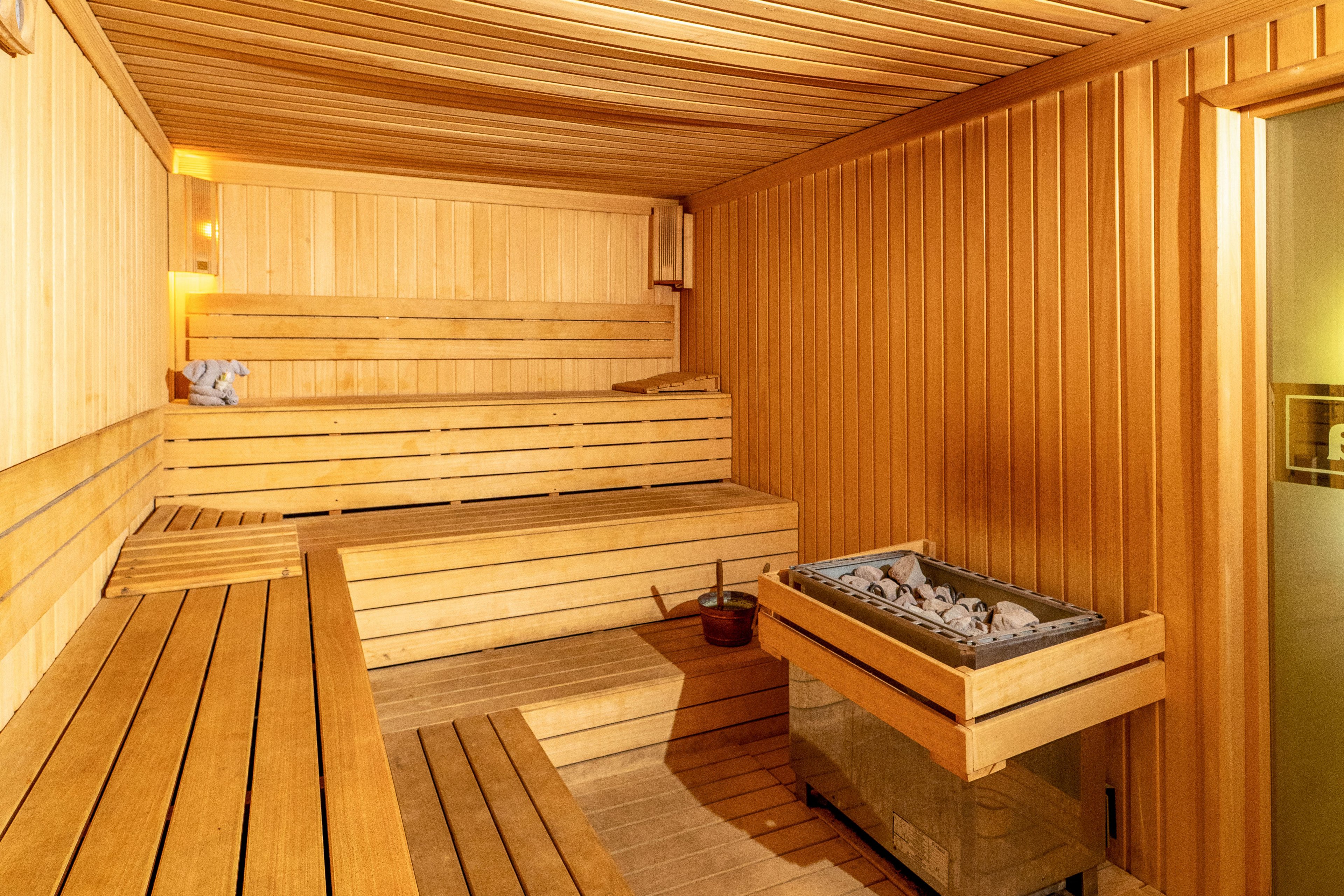 A warm, empty wooden sauna interior is shown with multiple tiered benches along the walls. The space is lined with smooth vertical and horizontal wooden panels, creating a cohesive natural look. A metal sauna heater filled with stones sits in the foreground, with a wooden bucket and ladle nearby. Soft lighting enhances the cozy, relaxing atmosphere of the room.