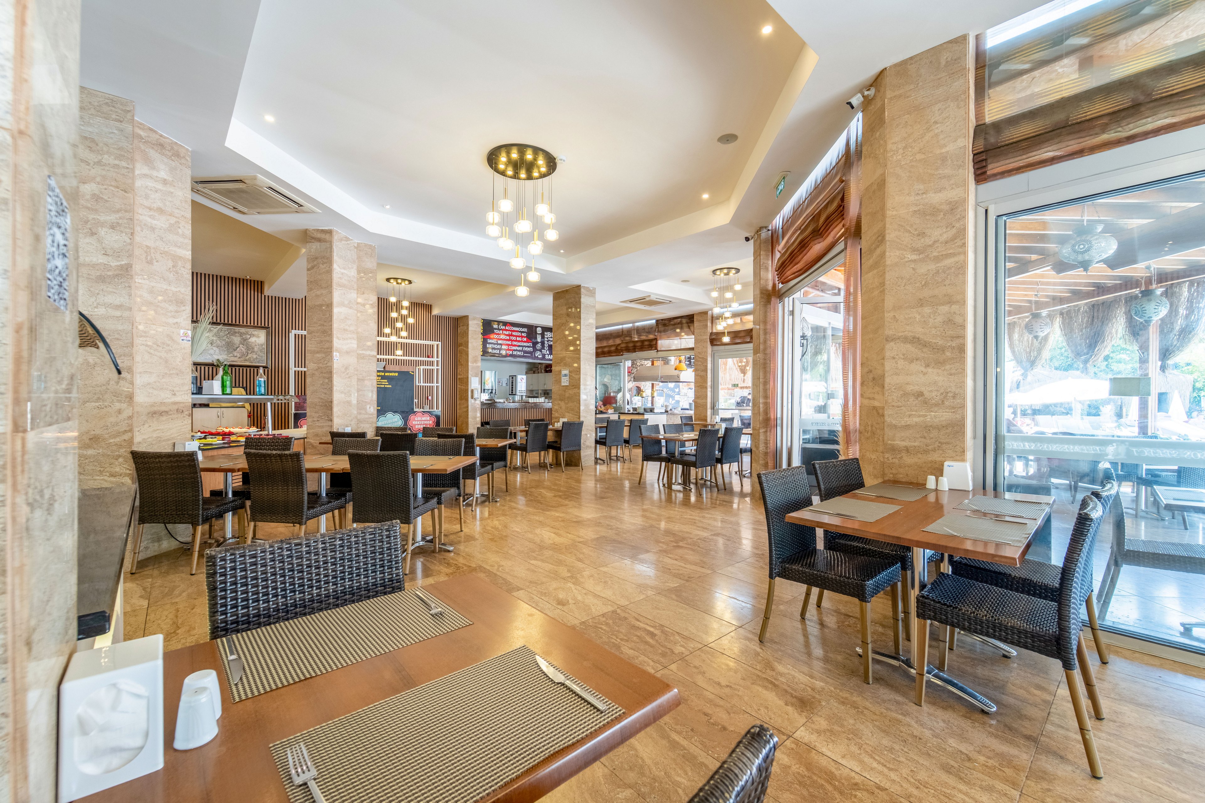 A spacious, contemporary restaurant interior features neatly arranged tables and wicker-style chairs on a polished floor. Place settings with cutlery and placemats are prepared, but the dining area is currently empty of guests. Large windows and glass doors let in abundant natural light, revealing an outdoor seating area beyond. Warm neutral tones, decorative lighting fixtures, and clean lines create an inviting, modern atmosphere suitable for casual dining.