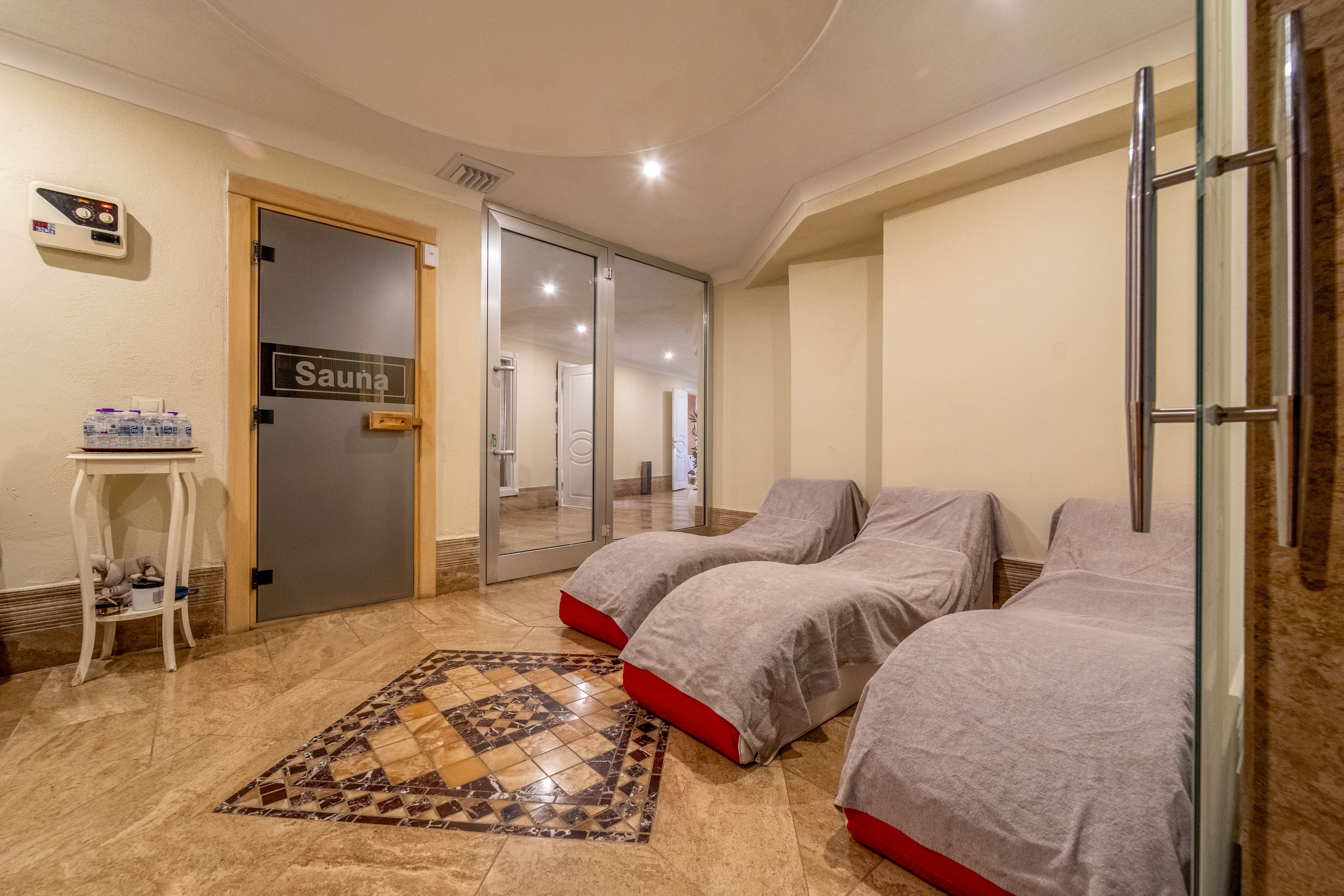 A spa relaxation room features several cushioned loungers covered with gray fabric and red bases arranged along a tiled floor. A glass door labeled "Sauna" leads to an adjacent sauna area, enhancing the wellness atmosphere. Warm beige walls, recessed lighting, and decorative floor tiles create a cozy, inviting setting. A small side table with water bottles and cups suggests a space designed for rest and hydration between sauna sessions.