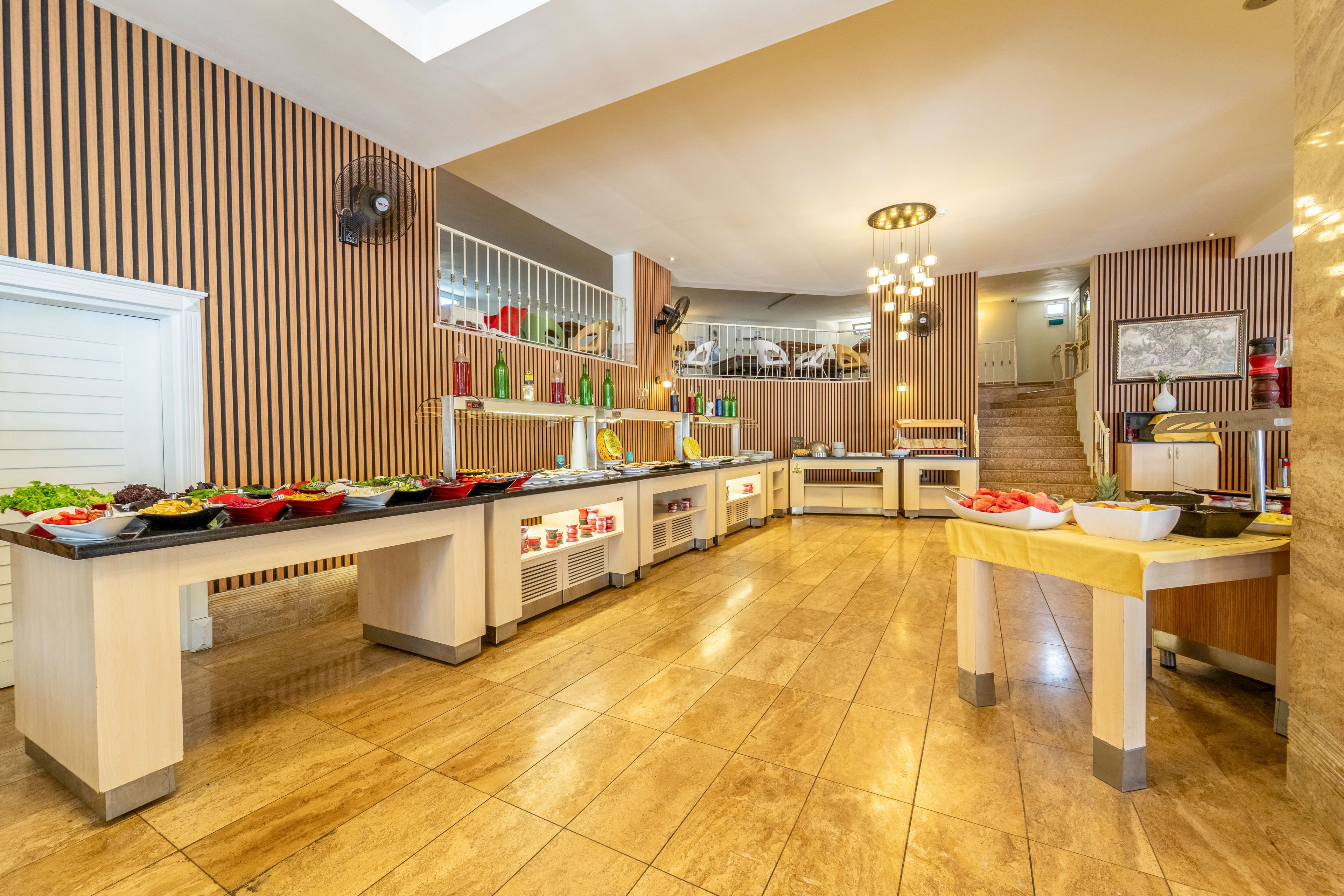 A spacious self-service buffet area is set up with various bowls and trays of fresh food, including salads and vegetables. Long light-colored counters with dark tops run through the room, offering multiple serving stations. The interior features warm wooden wall panels, tiled flooring, and modern ceiling lights that create a welcoming atmosphere. Shelves with bottles and decorative items line the back wall, and stairs lead up to an additional seating or service level.