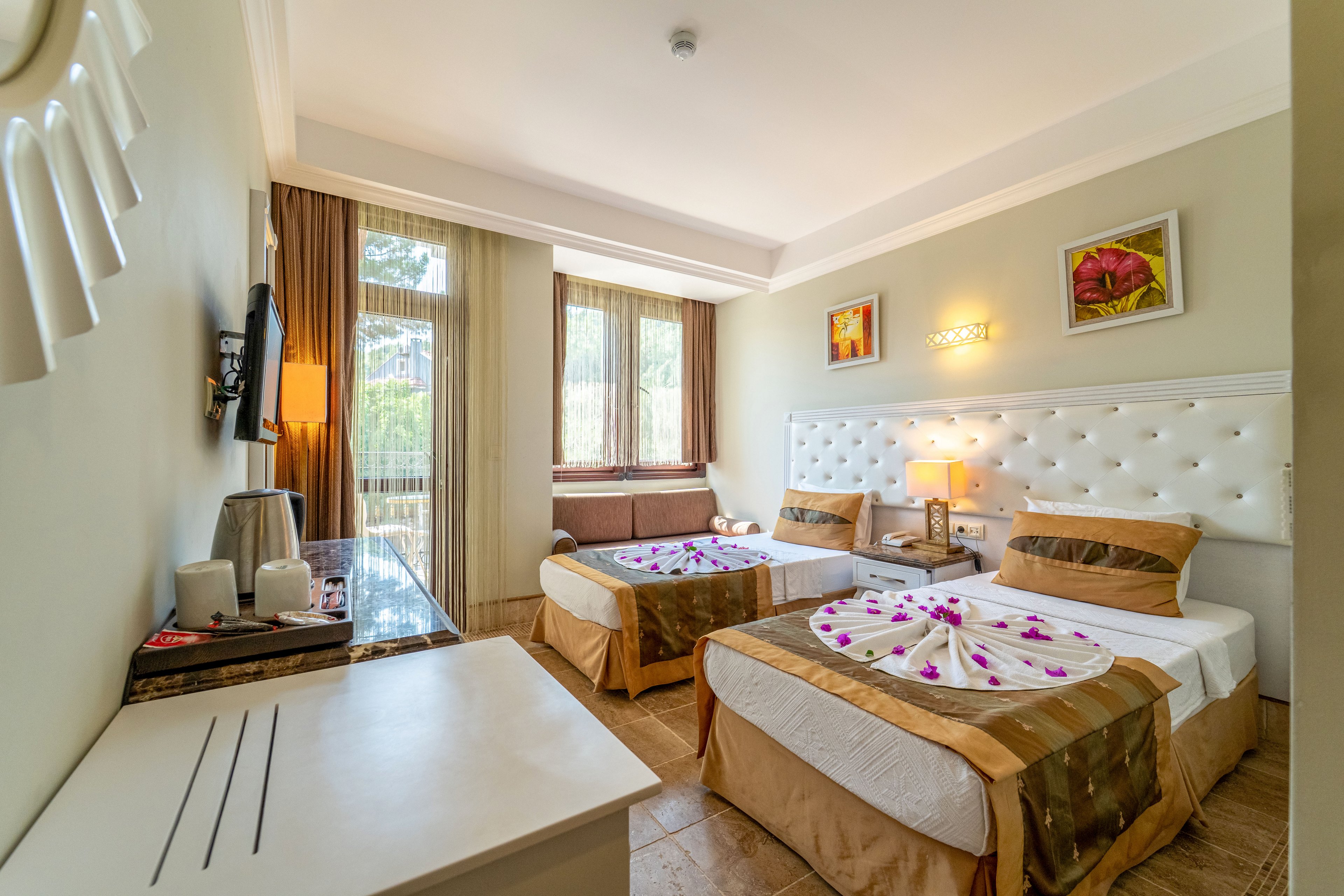 A well-lit hotel room features two neatly made twin beds with decorative folded towels and flower petals. Large windows with sheer curtains allow natural light to fill the space and offer a view of greenery outside. A white padded headboard, bedside lamp, and wall art add a touch of elegance and warmth. A desk with a television and accessories completes the cozy, inviting interior.