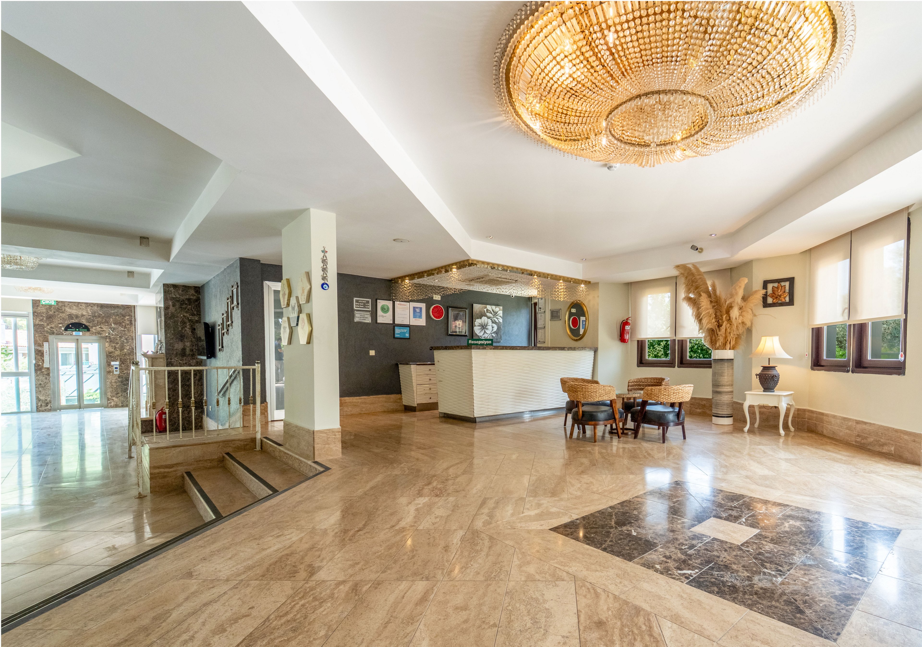 Spacious hotel lobby interior featuring a central reception desk and polished marble flooring. A large decorative chandelier hangs from the ceiling, illuminating the seating area with wicker chairs and a small table. Framed artwork and wall decor accent the neutral walls, while tall dried plants and vases add a natural touch. Windows along one side bring in daylight, and a staircase and glass entrance doors are visible in the background.