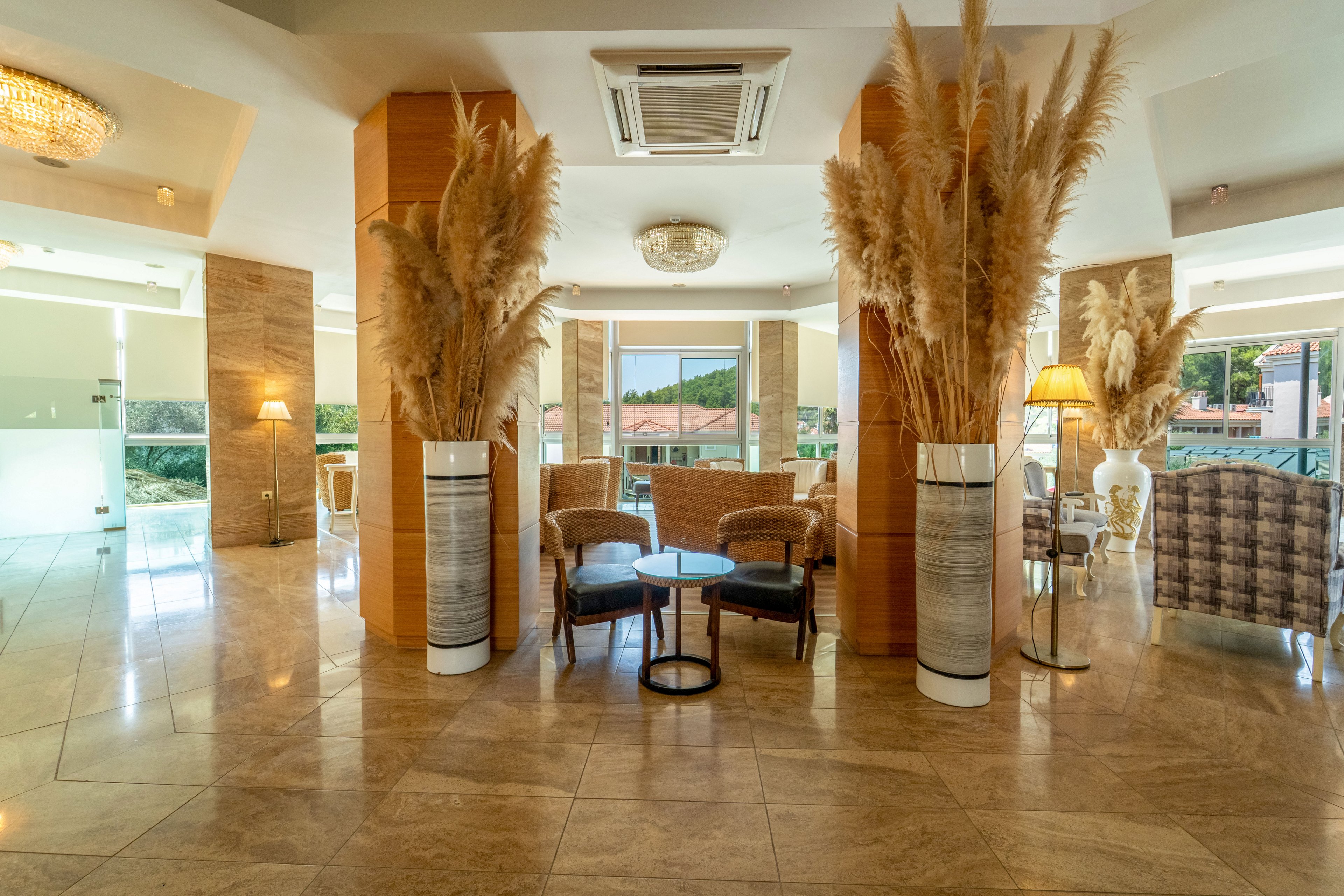 Spacious hotel lobby interior featuring tall vases filled with pampas grass as focal decor elements. The setting includes wicker armchairs around a small round table on a polished marble floor. Large windows in the background reveal a sunny outdoor view with rooftops and greenery. Warm lighting from ceiling fixtures and floor lamps creates an inviting, contemporary atmosphere.