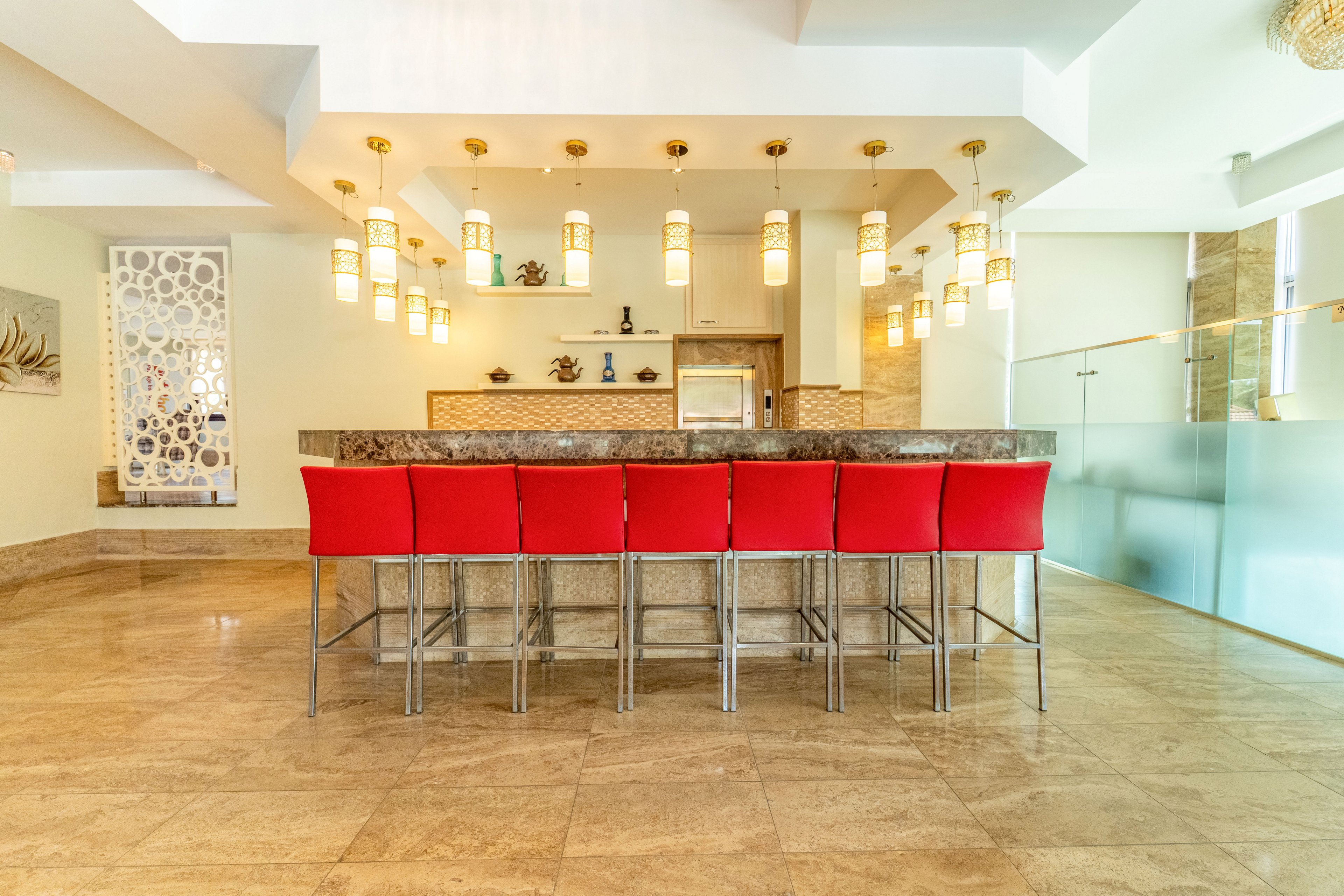 A contemporary bar area features a long stone countertop lined with bright red bar stools. Warm pendant lights hang from the ceiling, illuminating the polished floor and sleek finishes. Decorative wall elements and glass partitions add a modern, upscale touch to the open space. The overall scene feels clean, spacious, and inviting for social gatherings.