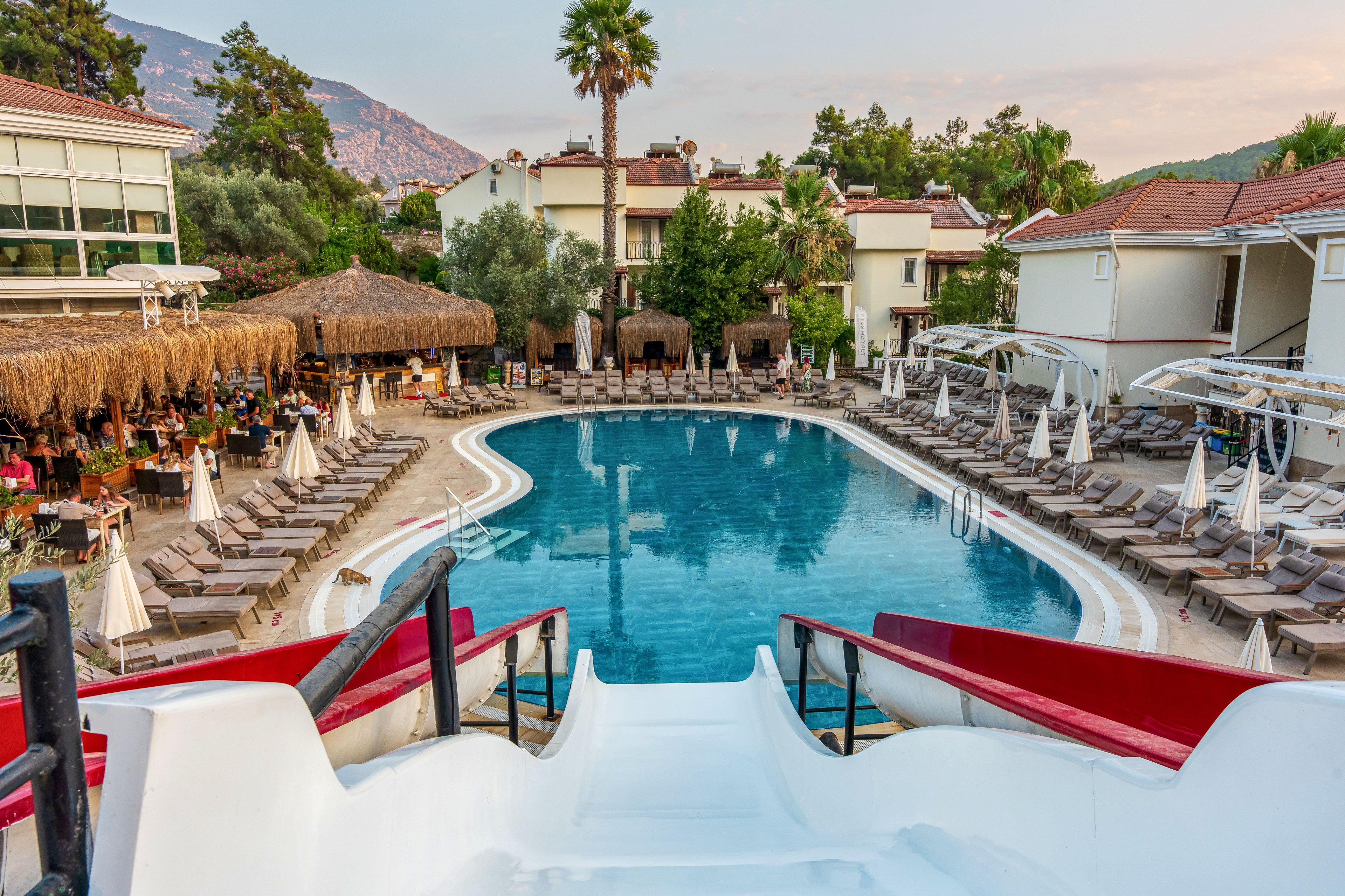 A large resort swimming pool is viewed from the top of a curved waterslide, with rows of sun loungers and umbrellas surrounding the water. Guests sit and relax at an open-air bar and dining area under thatched roofs on one side of the pool. Multi-story hotel buildings with balconies frame the scene, and tall palm trees rise above the courtyard. In the background, mountains and a soft evening sky create a tranquil vacation atmosphere.