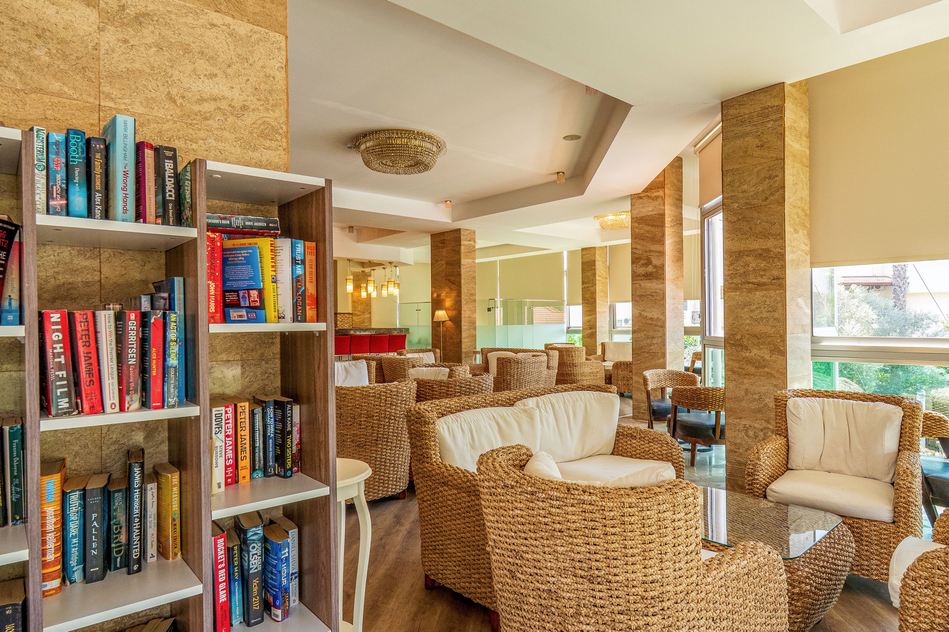 A bright lounge area features a tall bookshelf filled with colorful books beside a seating space. Wicker armchairs with white cushions are arranged around glass-topped tables, creating an inviting place to relax. Large windows and glass partitions let in natural light and offer views of greenery outside. Warm beige walls, wooden flooring, and elegant ceiling lights contribute to a comfortable, modern atmosphere.