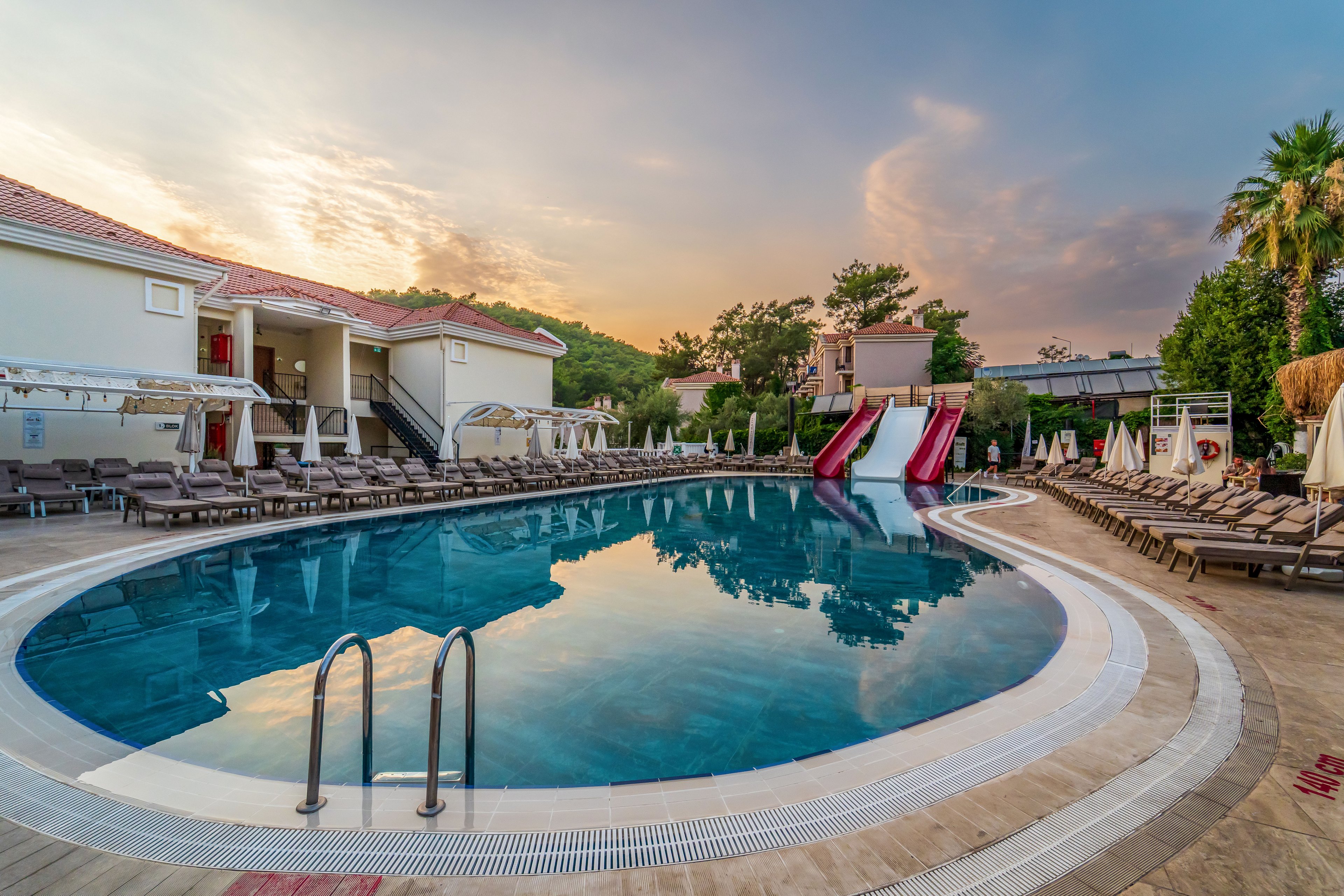 A large curved swimming pool is shown at a resort, surrounded by rows of sun loungers and umbrellas. Two colorful waterslides descend into the pool, adding a playful element to the tranquil setting. The scene is captured at sunset, with warm light reflecting on the calm water and soft clouds in the sky. Buildings and lush greenery frame the pool area, creating a relaxed vacation atmosphere.