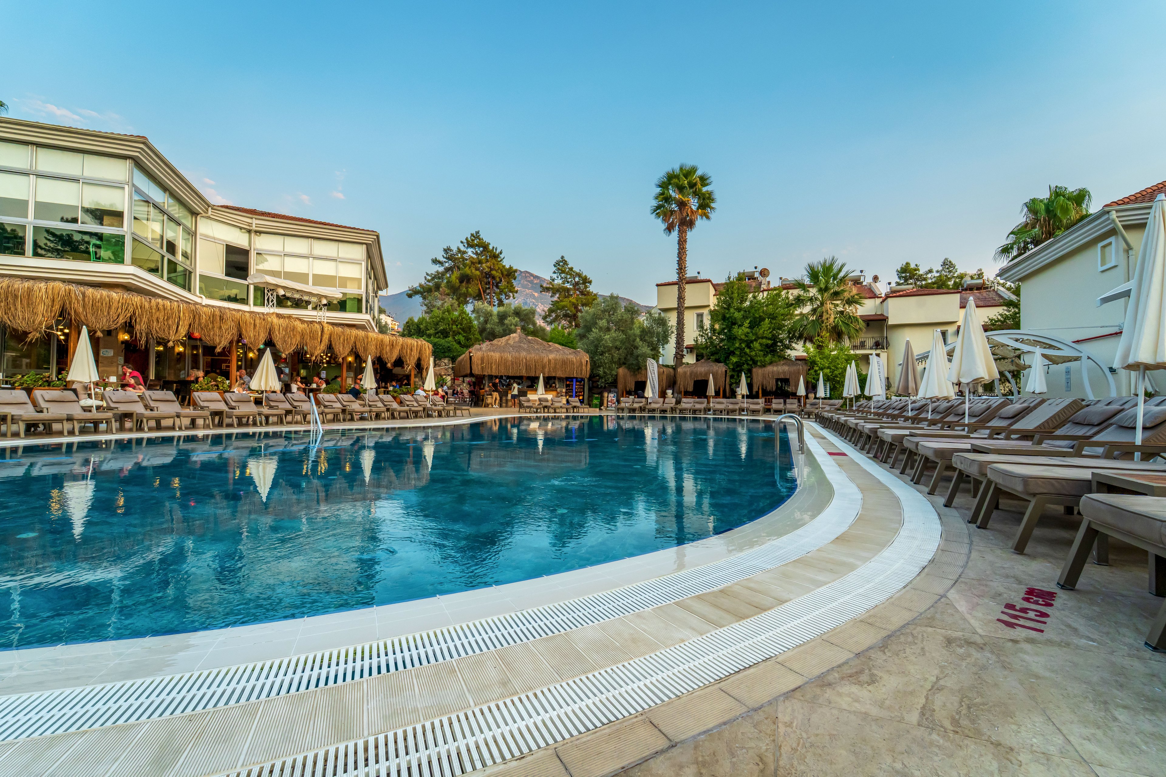 A curved resort swimming pool is surrounded by rows of empty sun loungers and white umbrellas. Modern hotel buildings and thatched-roof cabanas frame the water, with a tall palm tree rising above the scene. The clear blue pool reflects the sky, creating a calm and inviting atmosphere. Warm evening light suggests a tranquil vacation setting before guests arrive.