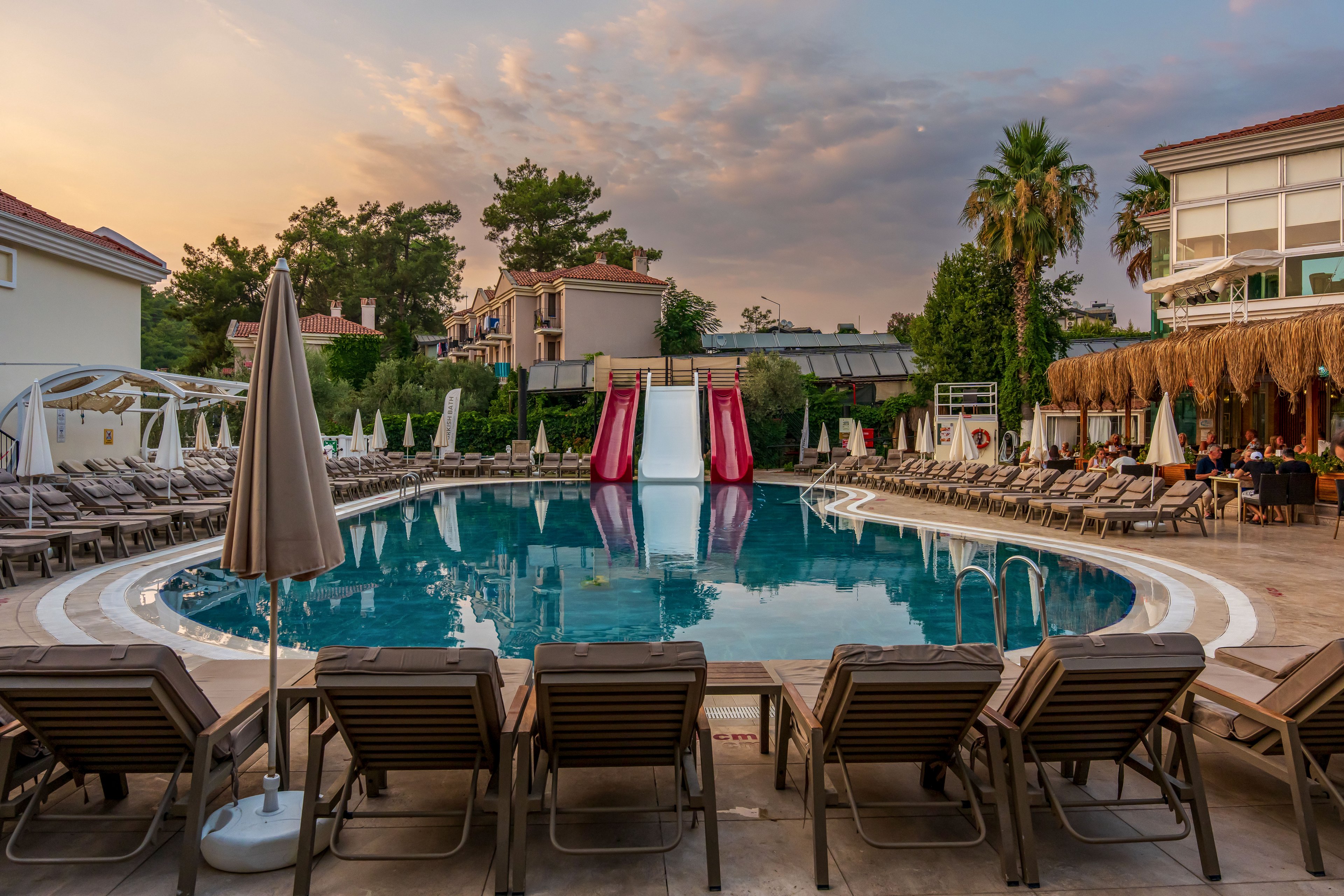 A circular resort swimming pool is surrounded by rows of empty lounge chairs and umbrellas at dusk. Two red waterslides descend into the pool, reflecting softly in the calm water. Guests sit and dine under a thatched-roof terrace on the right side, while low-rise buildings and tall trees frame the background. The sky is painted with warm evening colors, creating a relaxed vacation atmosphere.