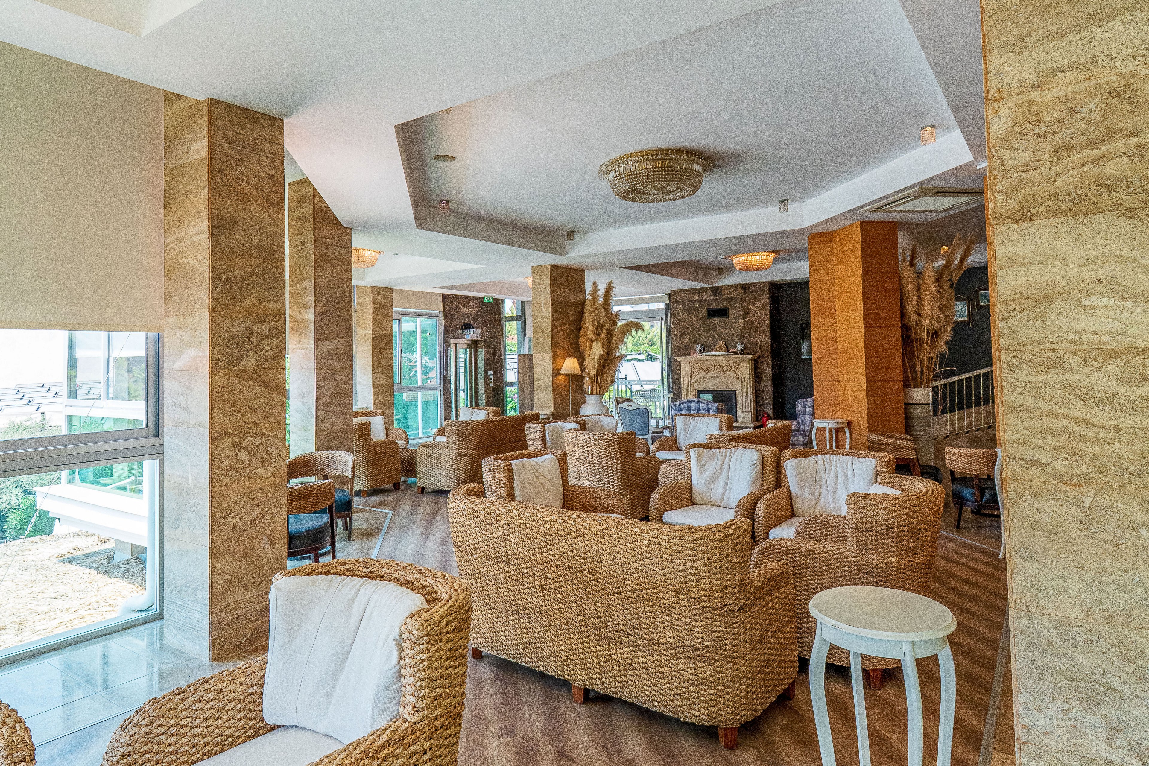 Spacious lounge area featuring multiple wicker armchairs and sofas with white cushions. Large windows bring in natural light and offer views of the outdoors. Warm stone columns, wooden flooring, and neutral decor create a cozy, upscale atmosphere. Decorative lighting and dried plants add to the relaxed, resort-like setting.