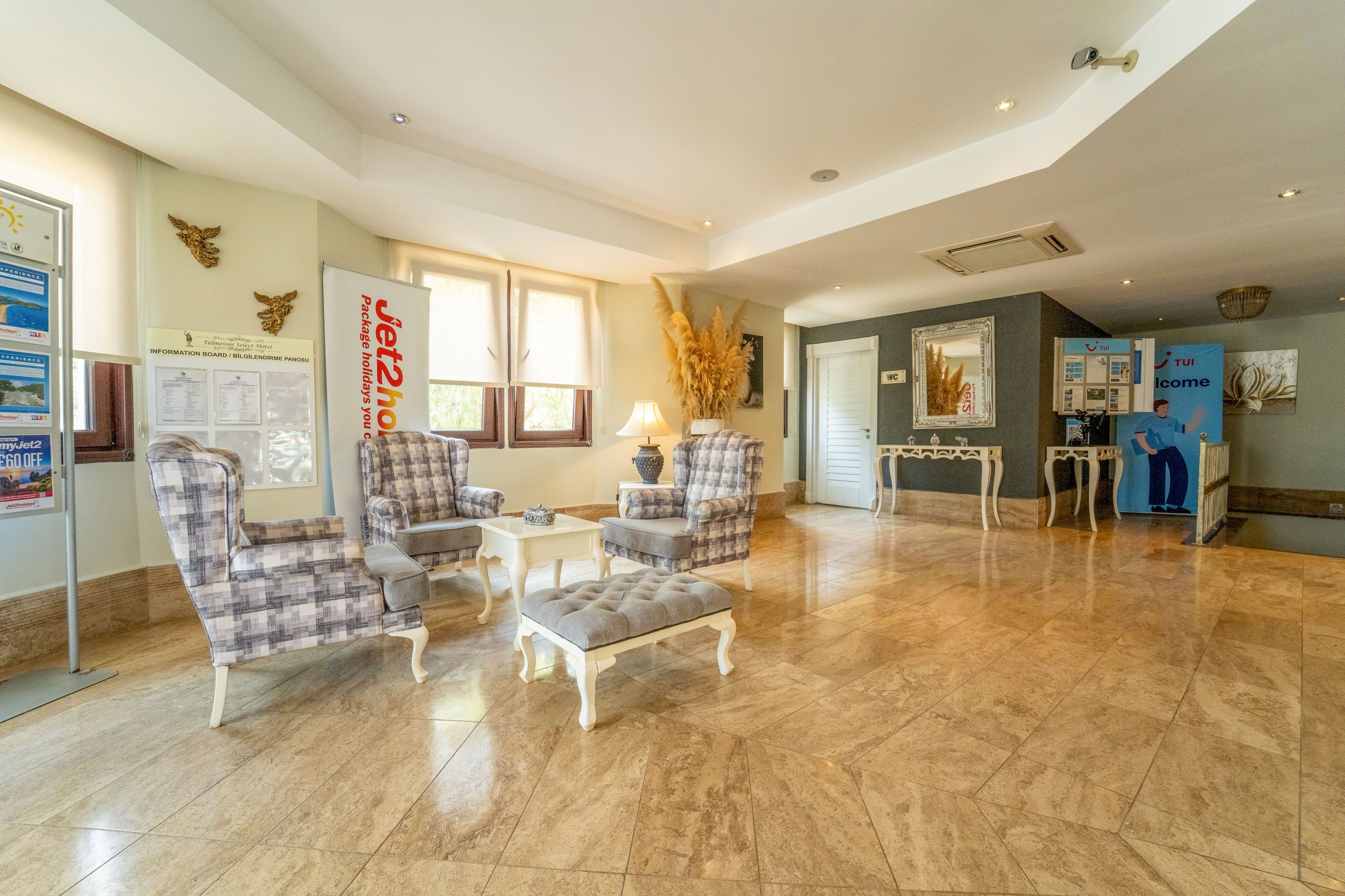 A spacious, sunlit lobby features four plaid armchairs arranged around a small cushioned ottoman on a polished marble floor. Large windows and recessed ceiling lights brighten the room, while decorative plants and wall art add warmth. A reception or information area with posters and a blue standing banner is visible in the background. The overall atmosphere is welcoming and contemporary, suitable for guests relaxing or waiting.