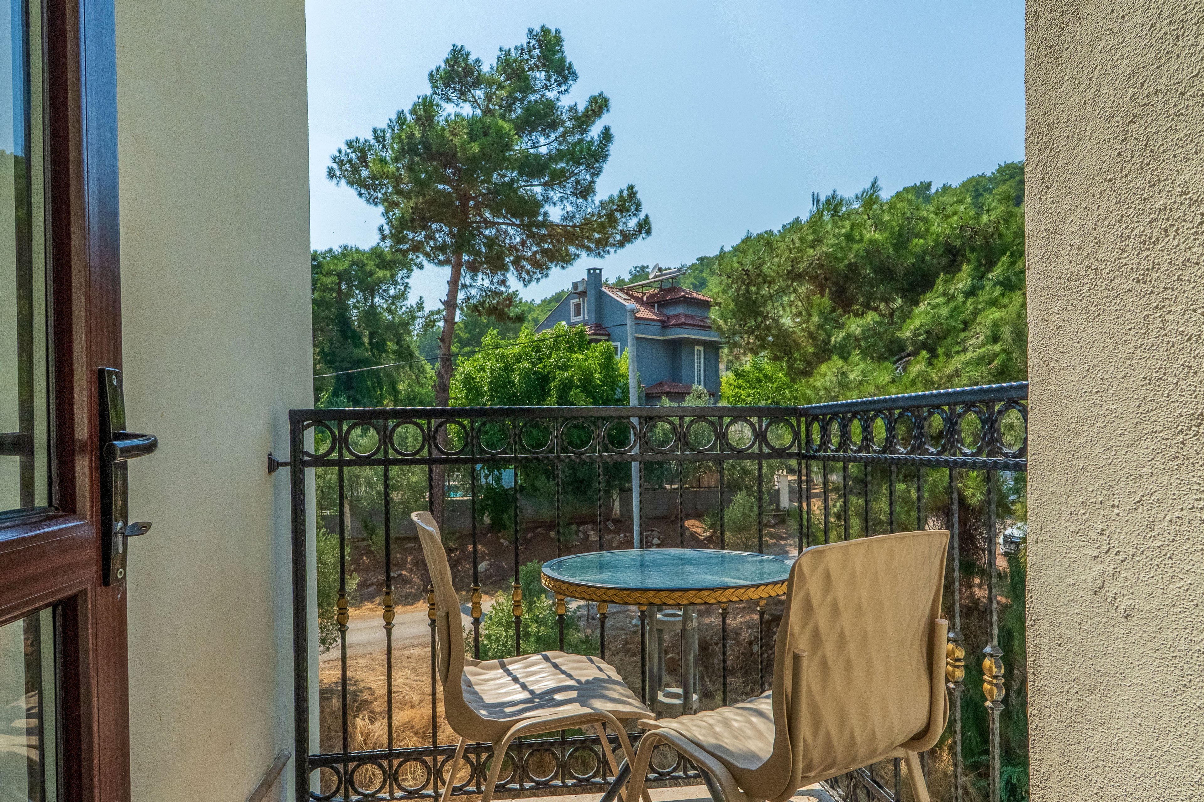 A small balcony features a round glass-top table and two modern chairs. Decorative metal railings frame the space, opening to a view of trees and a distant house. Bright sunlight creates strong shadows on the balcony floor, suggesting a warm day. The scene conveys a peaceful outdoor spot for relaxation or casual conversation.