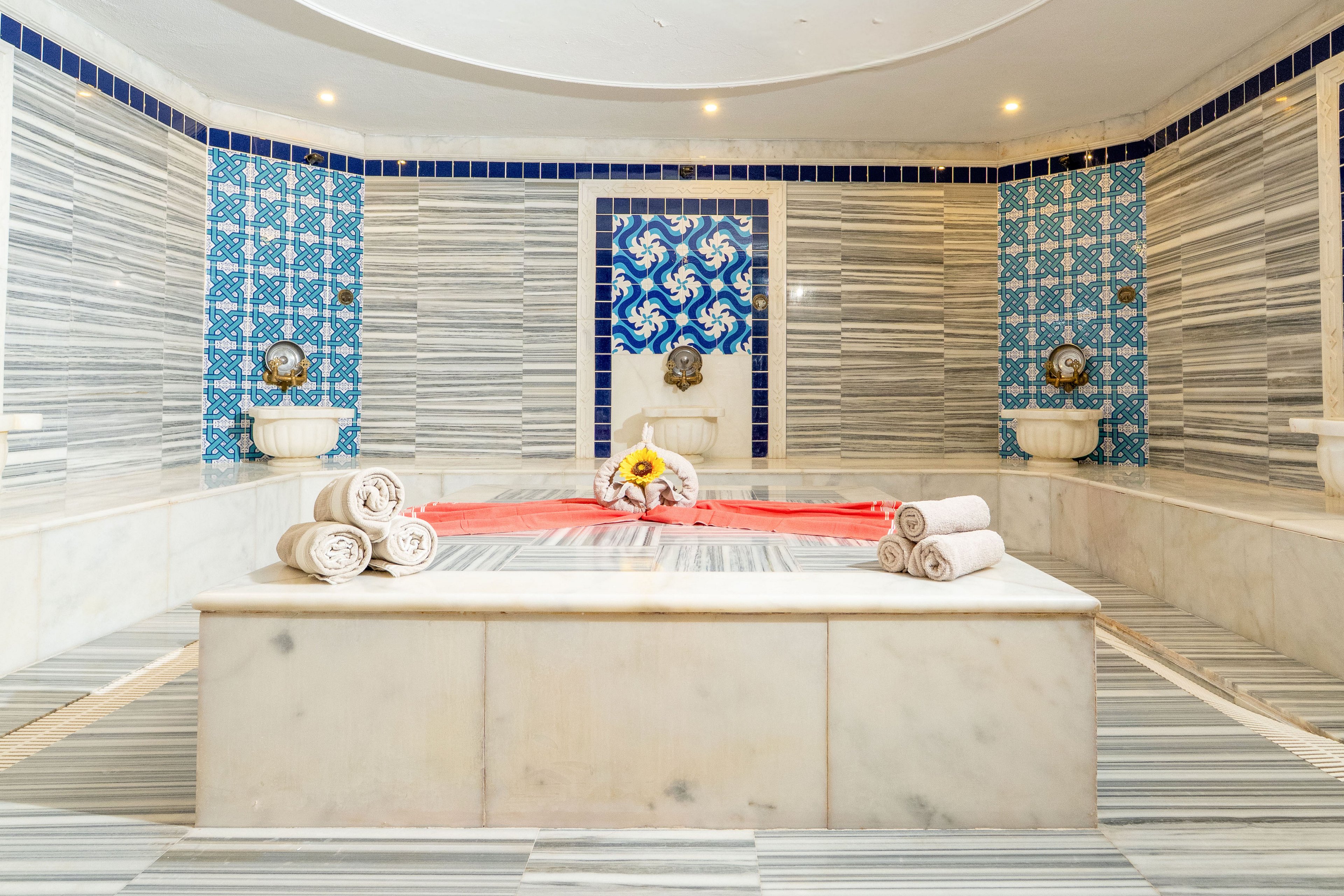 Elegant marble Turkish hammam with a central heated platform is prepared for a spa ritual. Rolled white towels and a red cloth are neatly arranged on the stone slab, suggesting relaxation and wellness treatments. Ornate blue and white patterned tiles frame traditional metal water basins on the walls. Soft ceiling lights create a warm, tranquil atmosphere in the pristine bathing space.