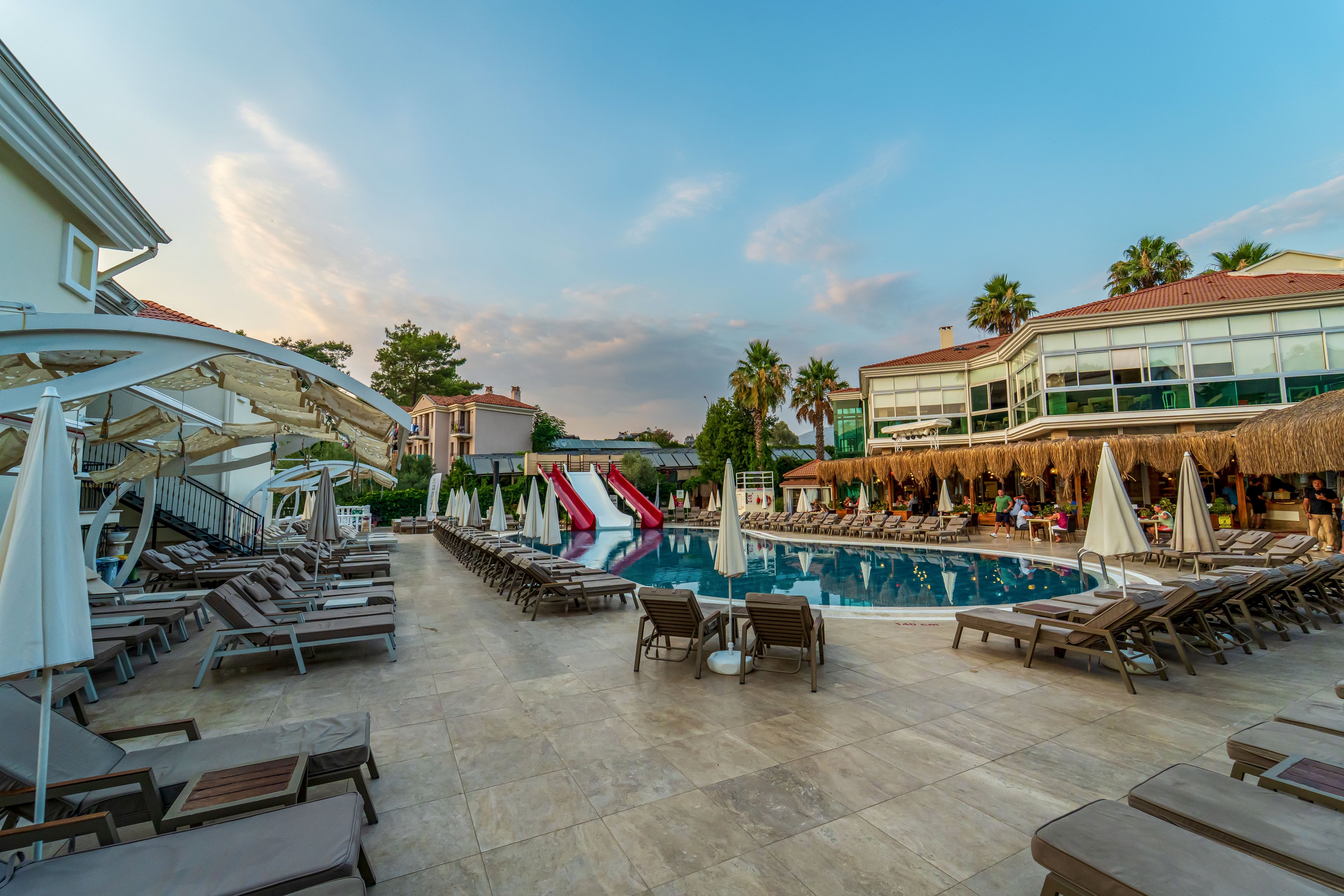 A large resort swimming pool is surrounded by rows of empty lounge chairs and umbrellas in the early evening light. A colorful waterslide and small play structures rise from one side of the pool, adding a sense of fun to the scene. Modern hotel buildings and palm trees frame the area, reflecting softly in the calm water. The sky above shows gentle clouds and warm tones, creating a relaxed vacation atmosphere.