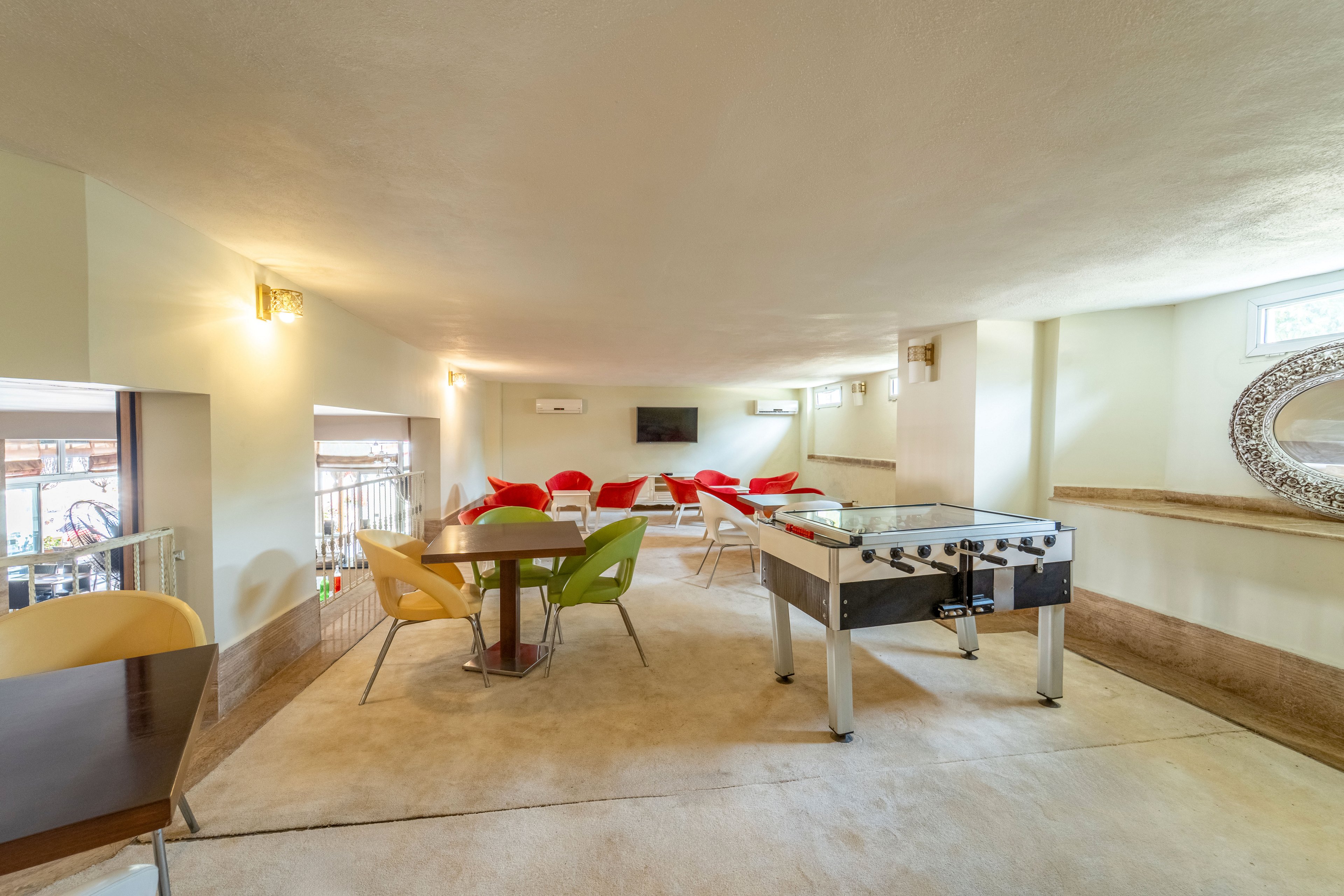 A spacious indoor recreation room features a foosball table as the central focus. Colorful chairs surround small tables, creating casual seating areas for socializing or games. The room is brightly lit with pale walls, a mounted television, and decorative elements including a large round mirror. Natural light enters through small windows, giving the space an airy, welcoming atmosphere.