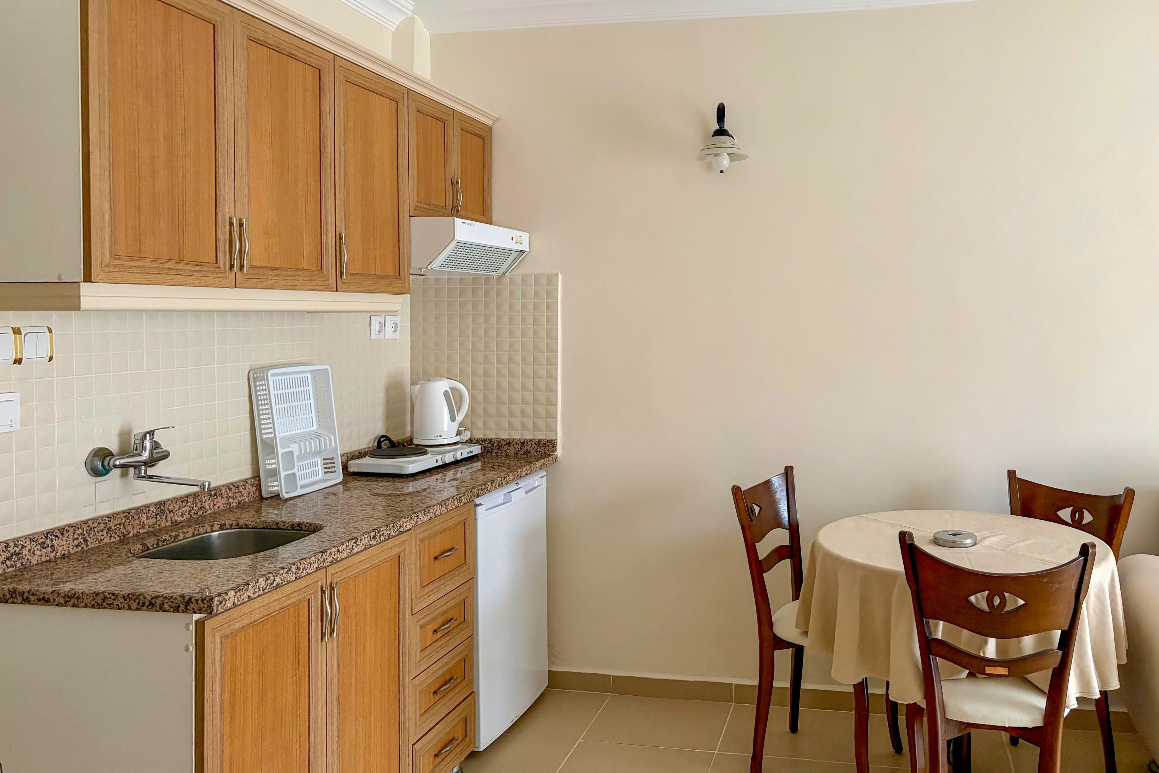 Compact kitchenette with wooden cabinets and a small sink is set against a light-colored tiled wall. A mini fridge, electric kettle, and single-burner cooktop sit on the granite-style countertop. To the right, a round table with a tablecloth and wooden chairs creates a simple dining area. Neutral walls and flooring give the space a clean, minimalist look suitable for a small apartment or vacation rental.