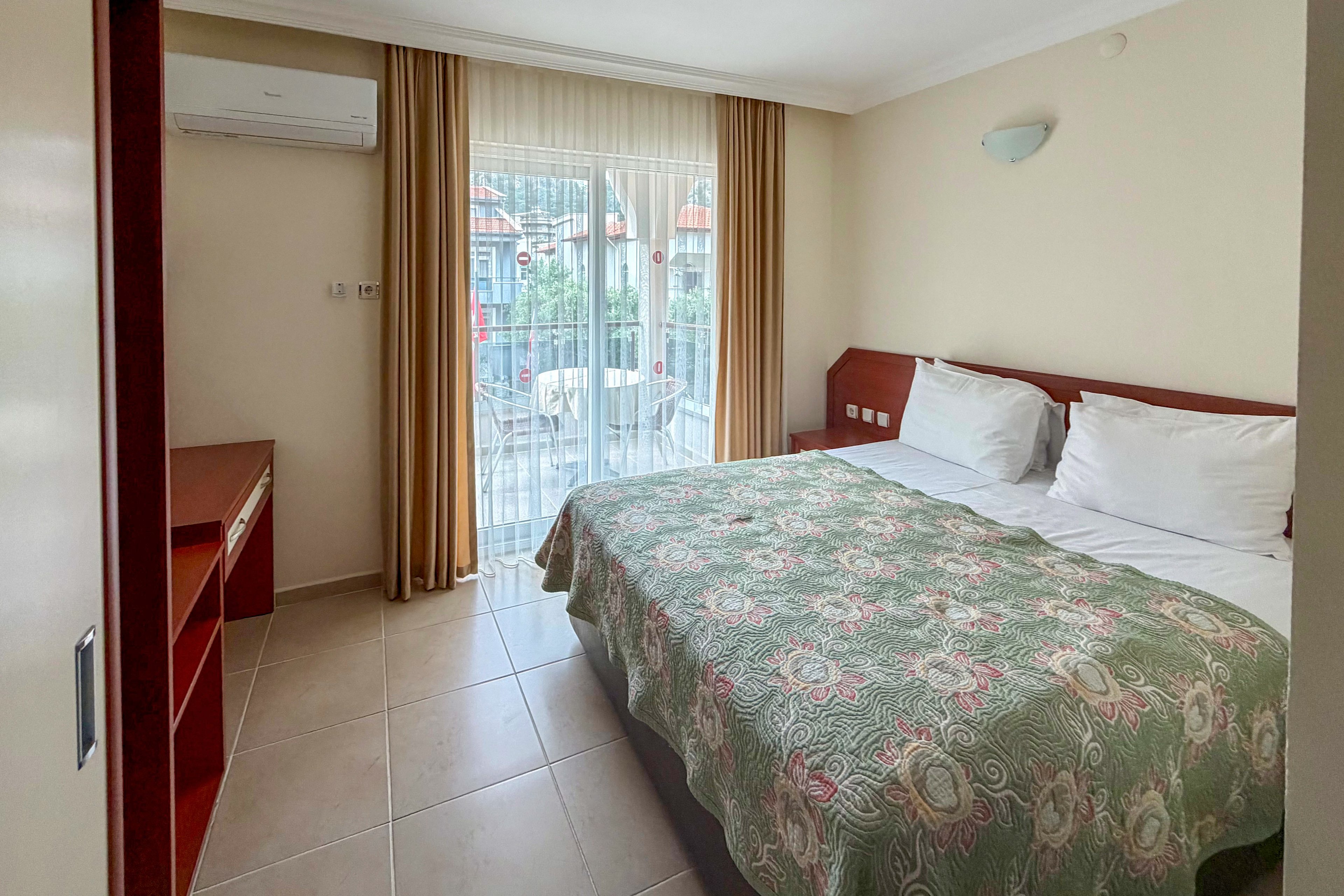 A tidy hotel bedroom features a double bed with a patterned green bedspread and white pillows. The room includes a wooden headboard, bedside table, and a small desk area opposite the bed. Large glass doors with beige curtains open to a balcony overlooking an outdoor area. Neutral walls and tiled floors create a clean, simple, and inviting atmosphere.