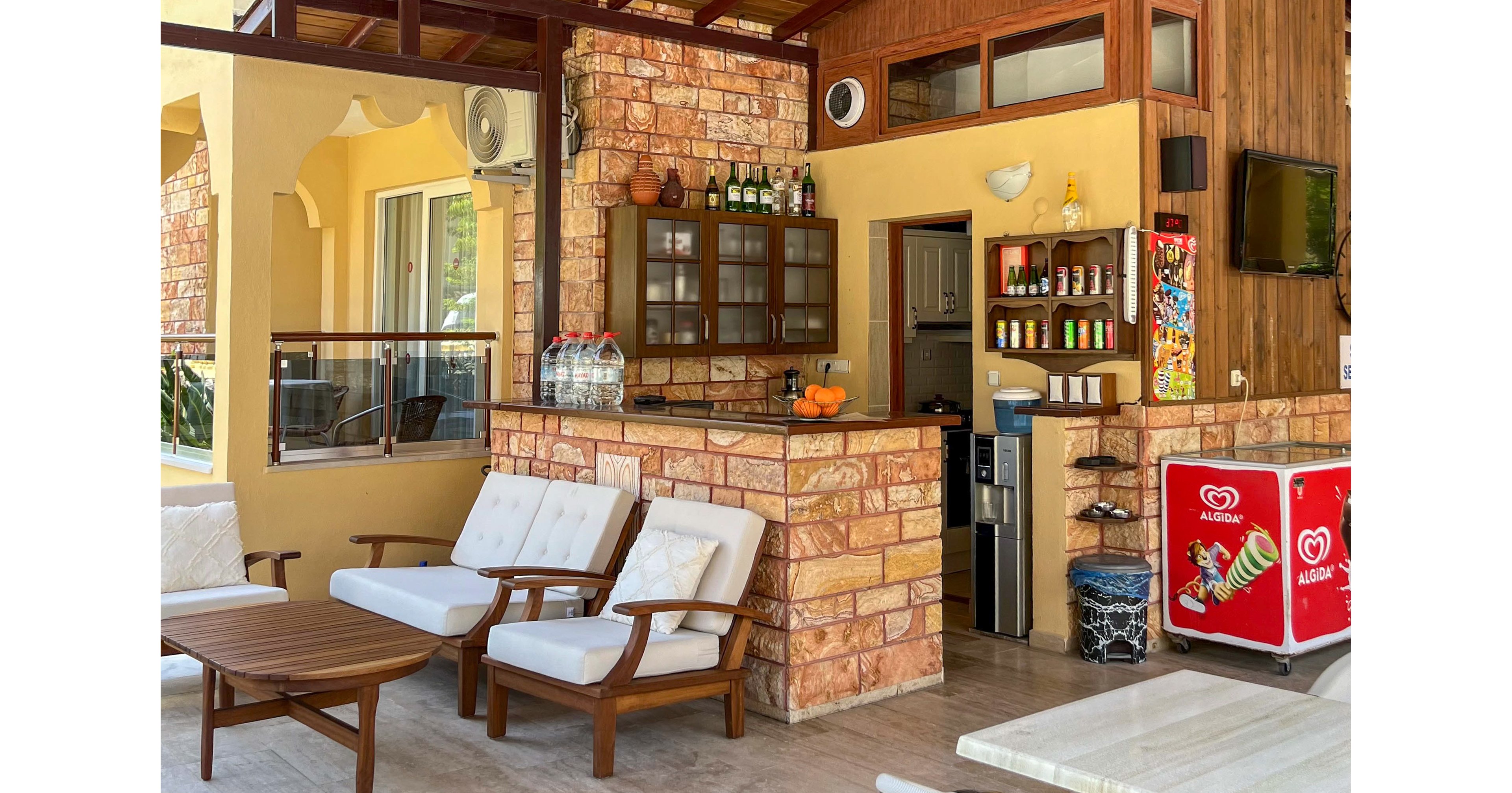 An outdoor snack bar features cushioned wooden chairs and a small table in a covered patio setting. The bar counter is built with stone and wood, displaying bottles, glasses, and a bowl of oranges. Refrigerated display units with ice cream graphics stand to the side, along with shelves of colorful packaged snacks and drinks. The warm yellow and brown tones, along with natural light, create a relaxed holiday atmosphere.
