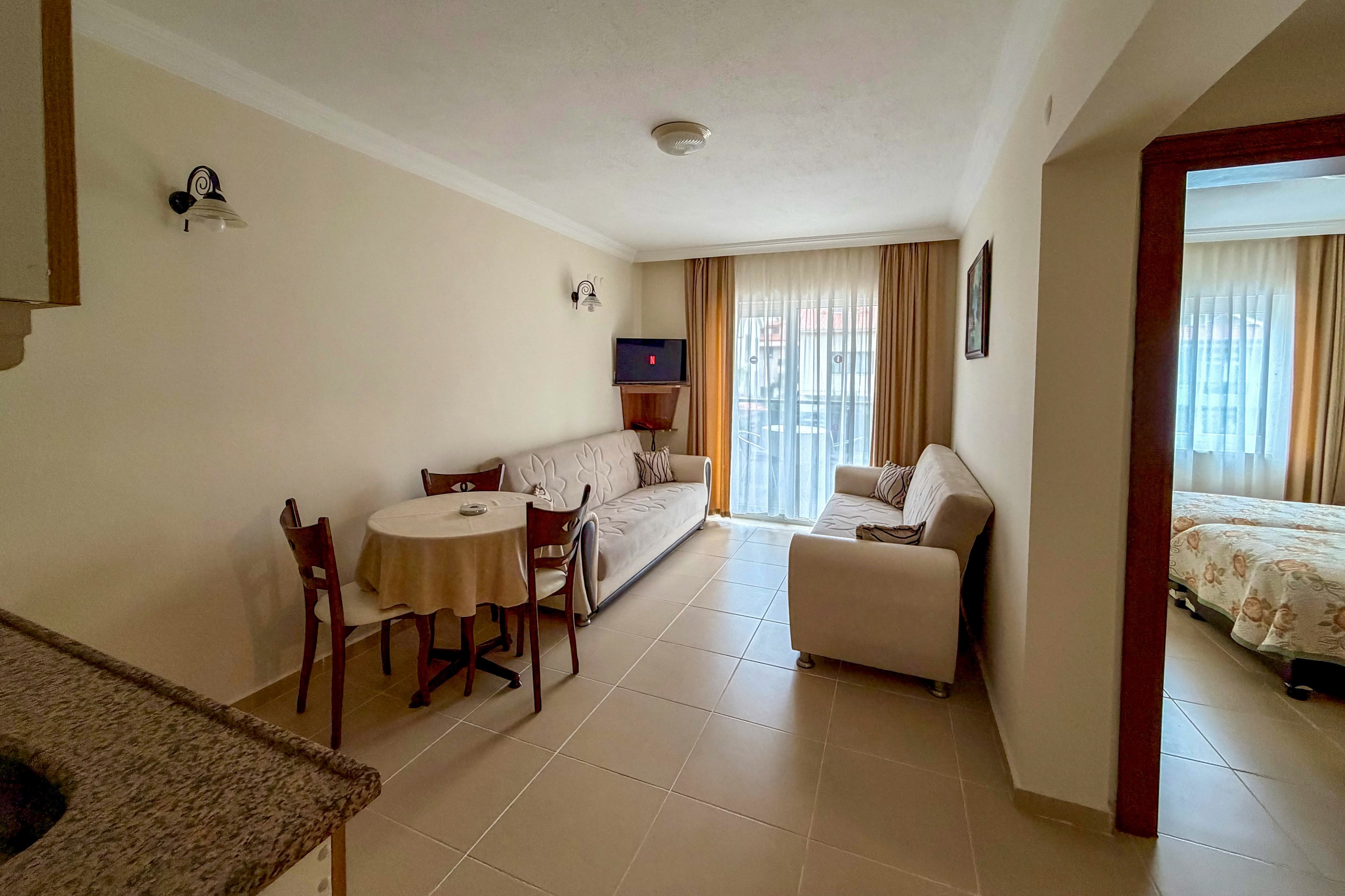 A compact hotel suite living area features a sofa, armchair, and small round dining table with wooden chairs. Large windows with sheer and beige curtains open to a balcony, filling the room with natural light. A wall-mounted television and simple wall sconces add functional comfort to the neutral-toned space. An adjacent bedroom with a patterned bedspread is visible through an open doorway, creating an inviting vacation or short-stay atmosphere.