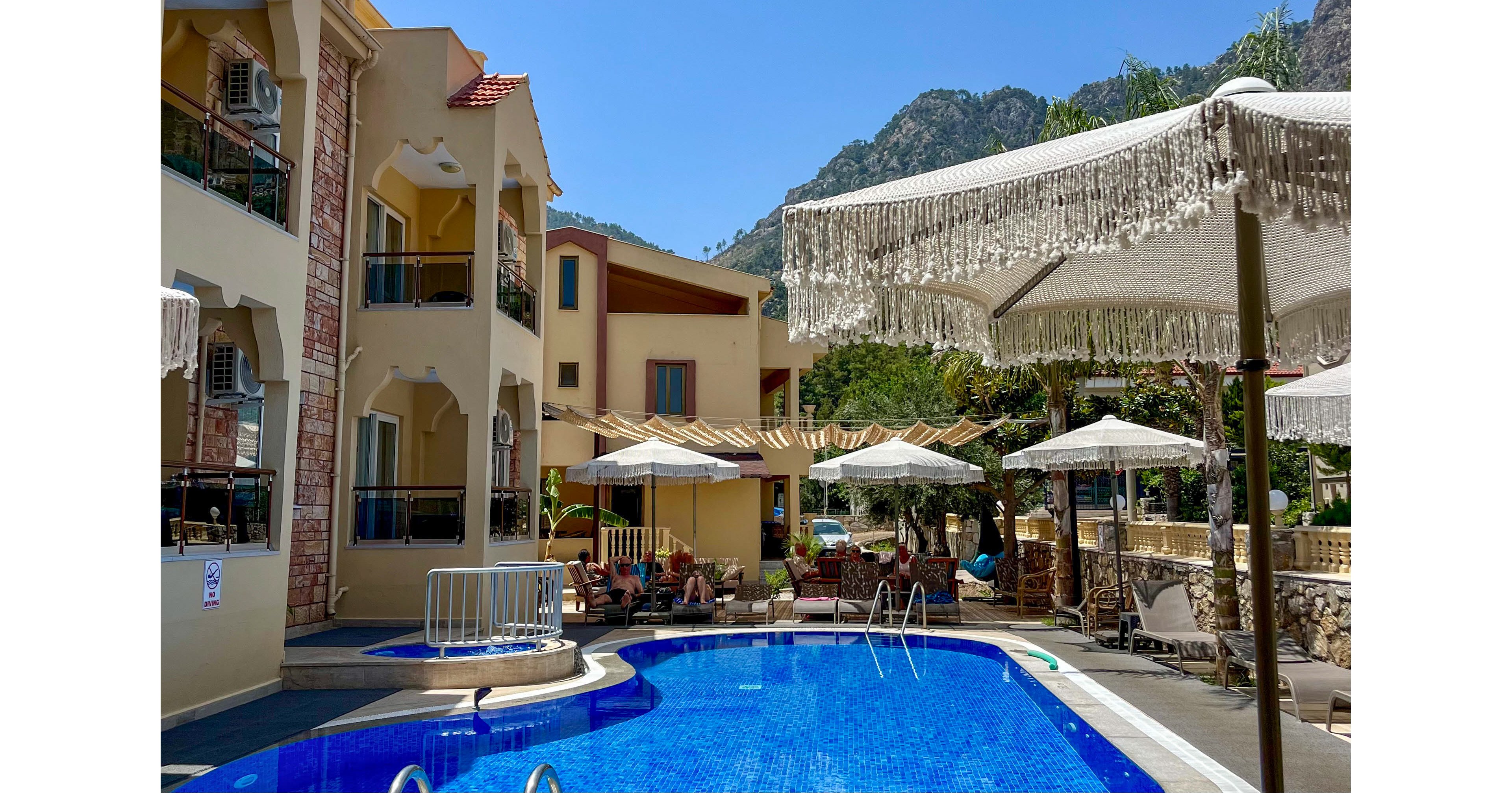 A bright hotel courtyard features a blue outdoor swimming pool surrounded by sun loungers and umbrellas. Guests relax in the shade of canopies and fringed parasols on a clear, sunny day. The multi-story building with balconies and air conditioning units frames the pool area. Green trees and rocky mountains rise in the background, creating a scenic vacation atmosphere.