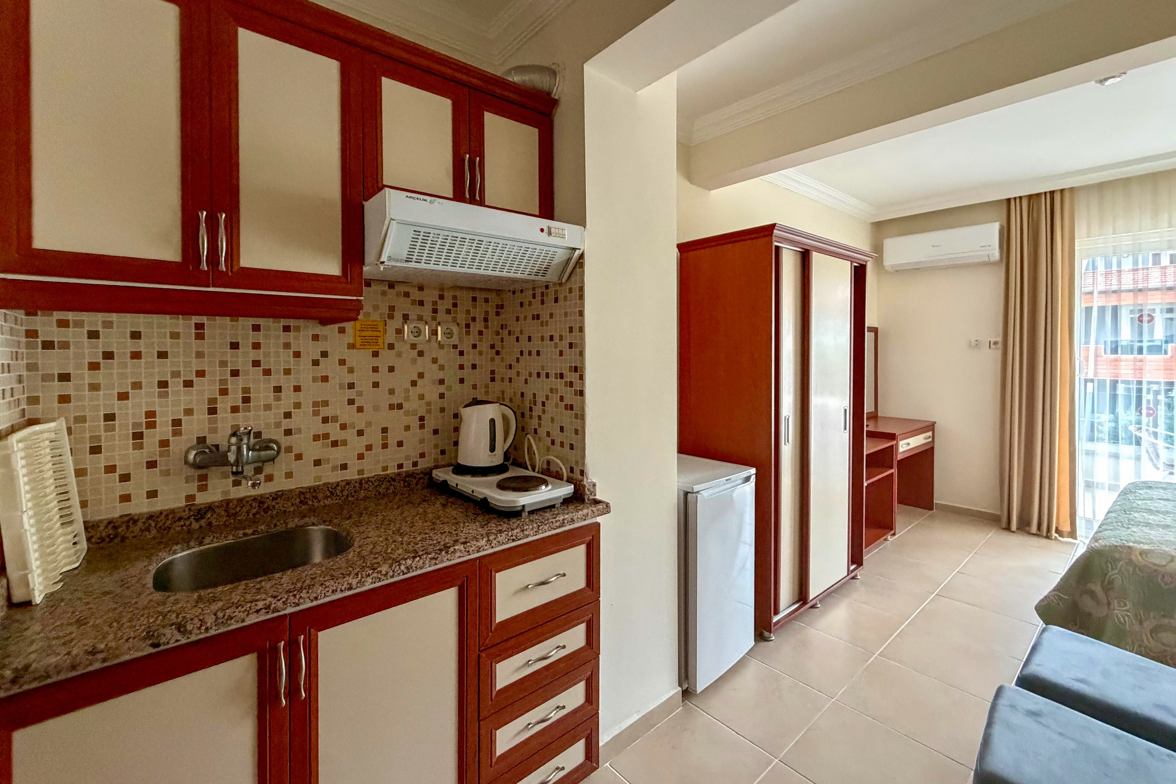 A compact studio apartment features a small kitchenette with a sink, stovetop, and mosaic tile backsplash. A kettle and dish rack sit on the granite countertop beneath upper cabinets and a range hood. Beyond the kitchen area, a mini refrigerator, wardrobe, and bed are visible in the open-plan room. Large windows with sheer curtains let in natural light and offer a view of the balcony and surrounding buildings.