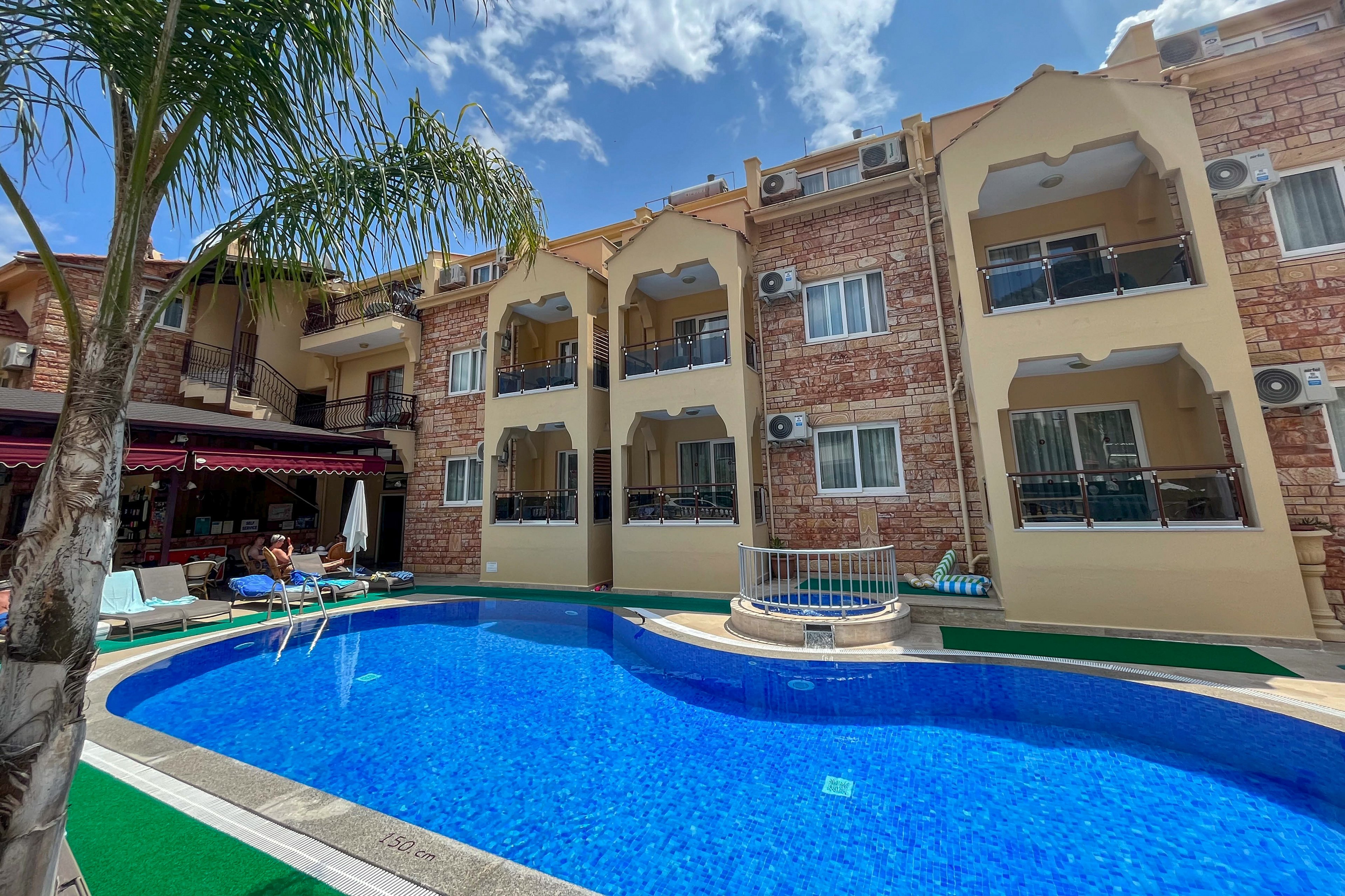 A bright hotel courtyard features a curved blue swimming pool in the foreground. Three-story buildings with balconies and stone facades surround the pool area. Lounge chairs, umbrellas, and a small seating area create a relaxed vacation atmosphere. A tall palm tree and a partly cloudy blue sky emphasize the warm, sunny weather.