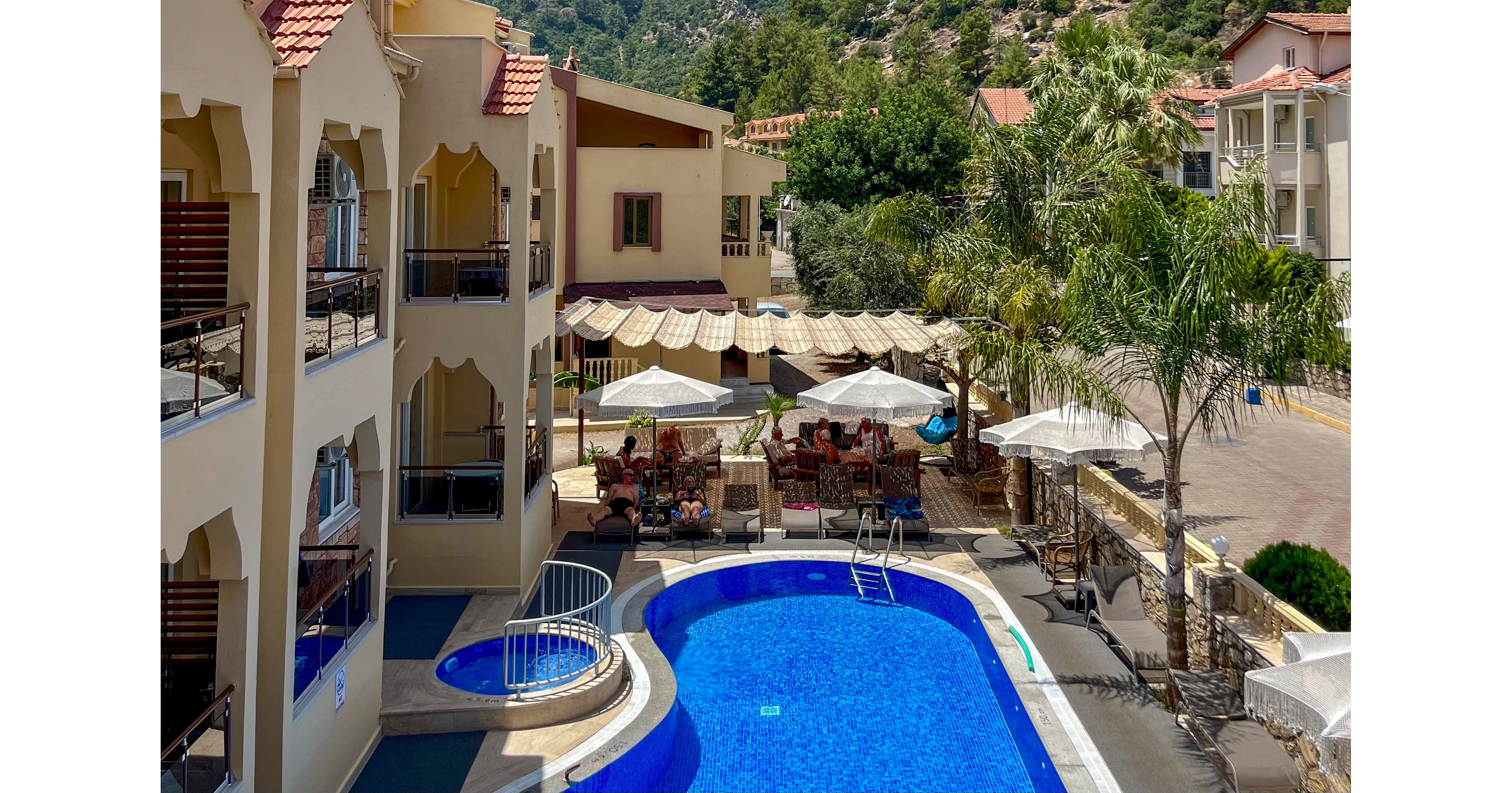 A bright hotel courtyard features a curved blue swimming pool with an attached circular hot tub. Guests relax under large white umbrellas near an outdoor café area beside the pool. Mediterranean-style buildings with balconies and red-tiled roofs surround the courtyard. In the background, green forested hills and mountains rise under a clear, sunny sky, creating a warm vacation atmosphere.