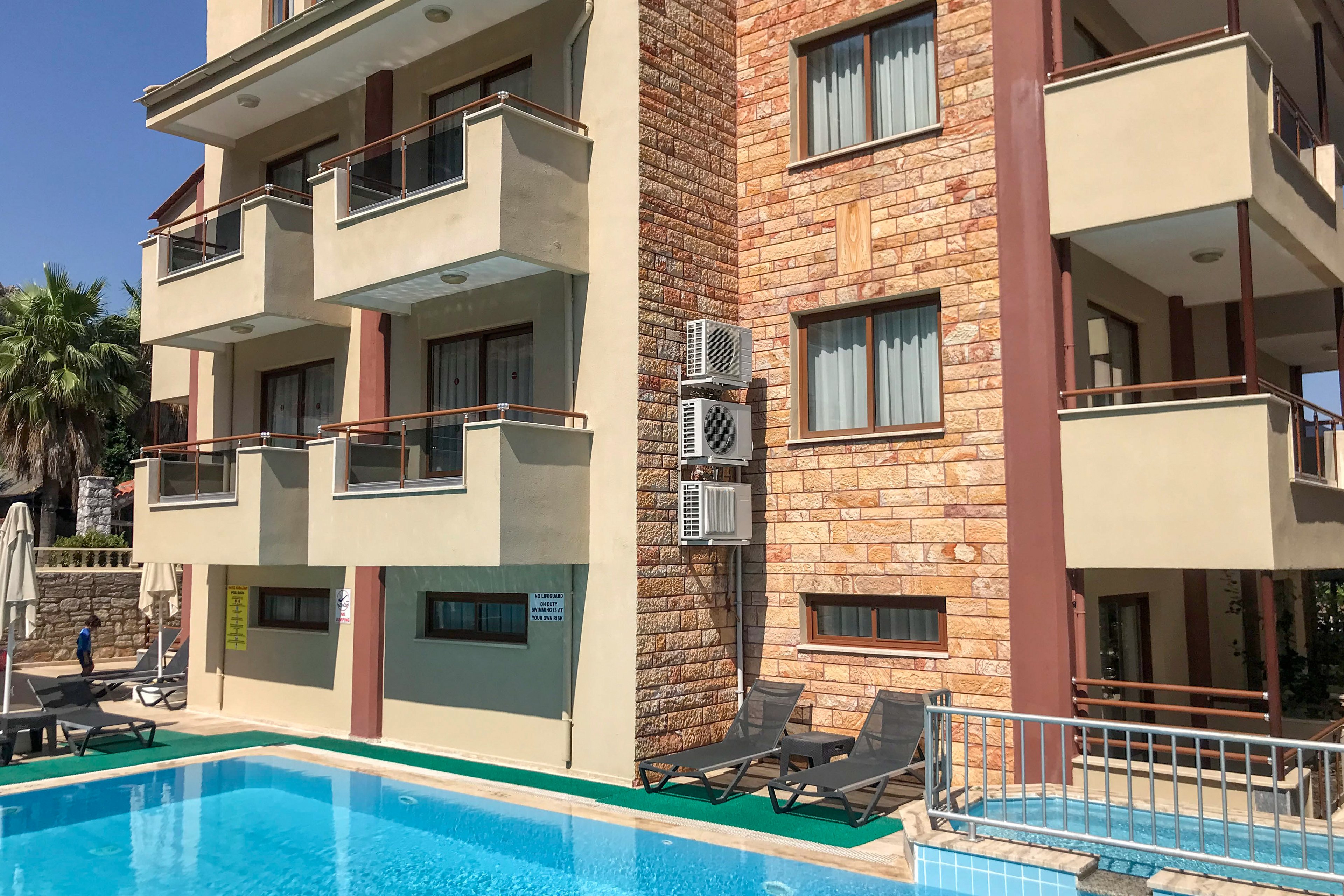 A modern multi-story apartment building stands beside a clear blue outdoor swimming pool. The structure features beige walls, stone cladding, and several balconies with metal railings. Poolside lounge chairs and a closed umbrella are arranged along the deck, suggesting a relaxing leisure area. Air conditioning units are mounted on the exterior wall, and palm trees are visible in the background under a bright, sunny sky.