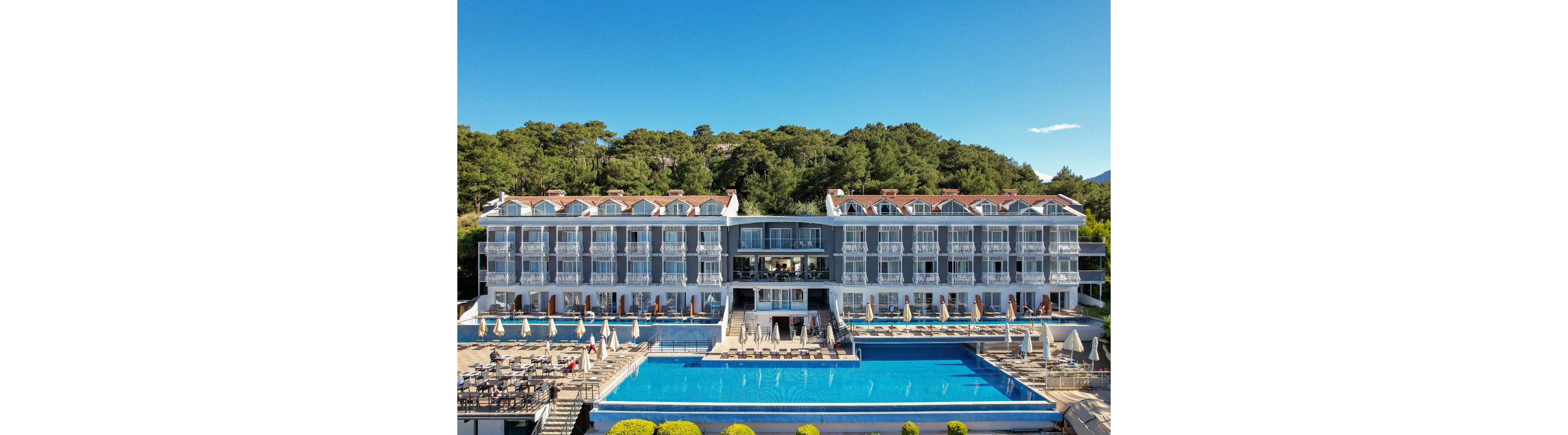A multi-story seaside resort hotel stands in front of a large blue infinity pool. Rows of balconies face the water, each with white railings and red-tiled roofs above. Sun loungers and umbrellas line the poolside terrace, creating a relaxed vacation atmosphere. Behind the building, a forested hillside rises under a clear blue sky. The scene suggests a tranquil coastal getaway with modern amenities and scenic surroundings.
