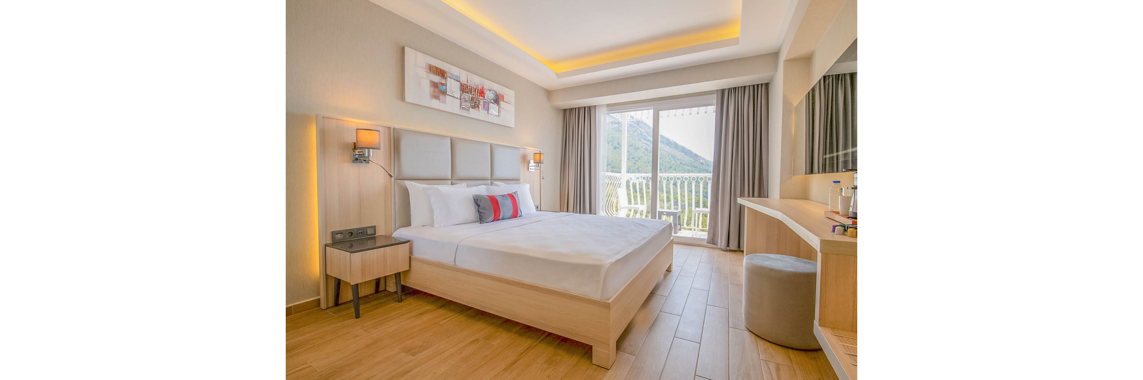 A modern hotel bedroom features a neatly made double bed with white linens and accent pillows. The room is decorated in light wood tones with soft, warm lighting and minimalist furniture including a bedside table, desk, and stool. Large glass doors open onto a balcony with a white railing, revealing a scenic mountain landscape outside. Neutral curtains, wall art above the bed, and a clean, uncluttered layout create a calm and inviting atmosphere.