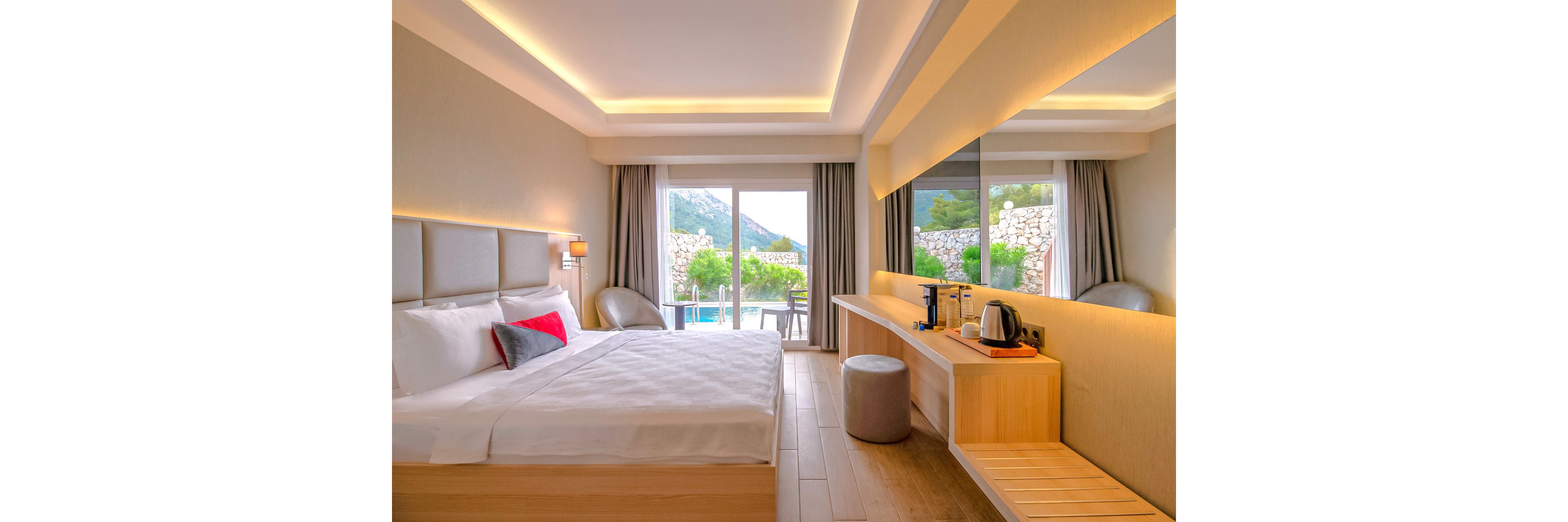 A contemporary hotel bedroom features a large neatly made bed with white linens and a small accent pillow. Floor-to-ceiling glass doors open to a patio with a pool and scenic mountain landscape in the background. A long wooden desk holds a kettle, cups, and a coffee station beside a large wall mirror. Soft recessed lighting and neutral tones create a calm, inviting atmosphere.