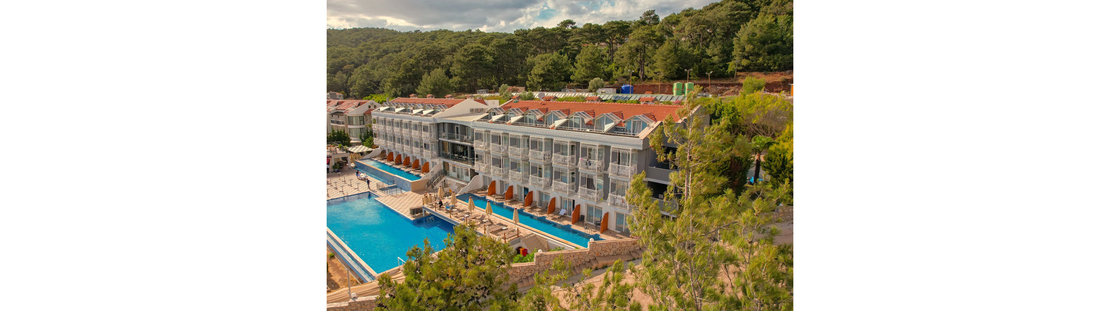 A contemporary multi-story resort complex is nestled on a forested hillside. The building features rows of balconies and glass-fronted rooms facing a large rectangular swimming pool. Sun loungers and umbrellas line the poolside, creating a relaxed vacation atmosphere. Dense green trees surround the property, contrasting with the blue water and the red-tiled roofs. The scene suggests a tranquil holiday destination in a natural setting.