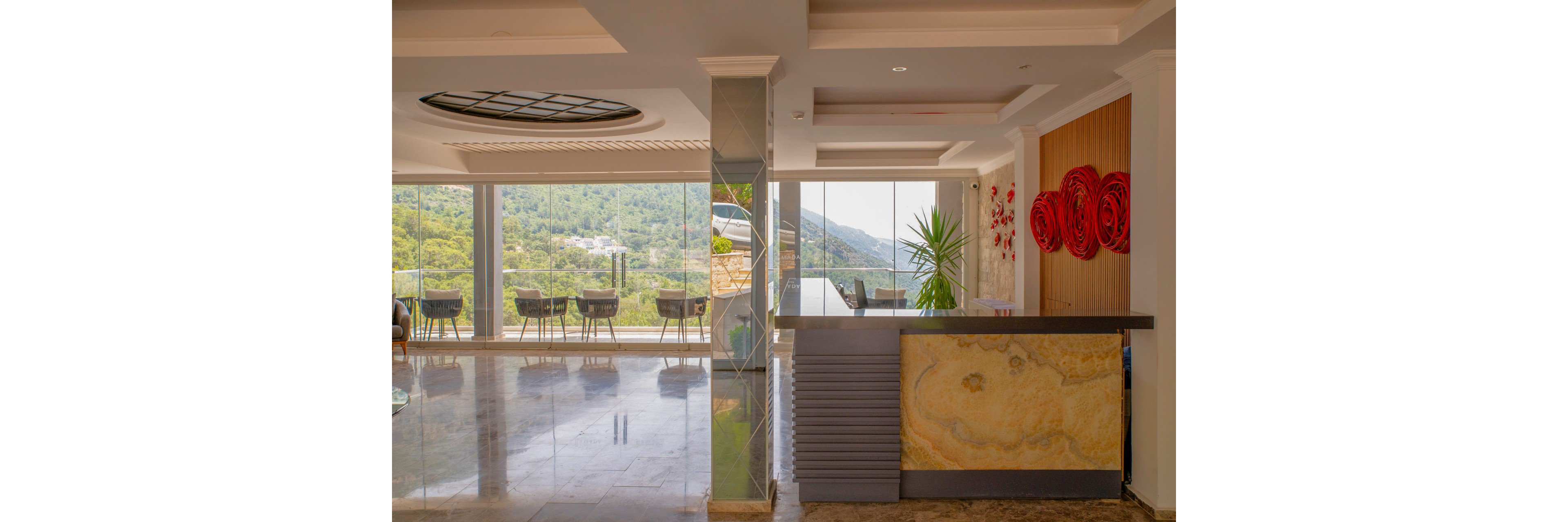 A contemporary hotel lobby features a sleek reception desk with a stone front panel. Large floor-to-ceiling windows open onto a terrace furnished with tables and chairs overlooking a lush, mountainous landscape. The interior includes polished floors, glass partitions, and a decorative ceiling light. Red circular wall art and a tall potted plant add color and contrast to the neutral-toned space.