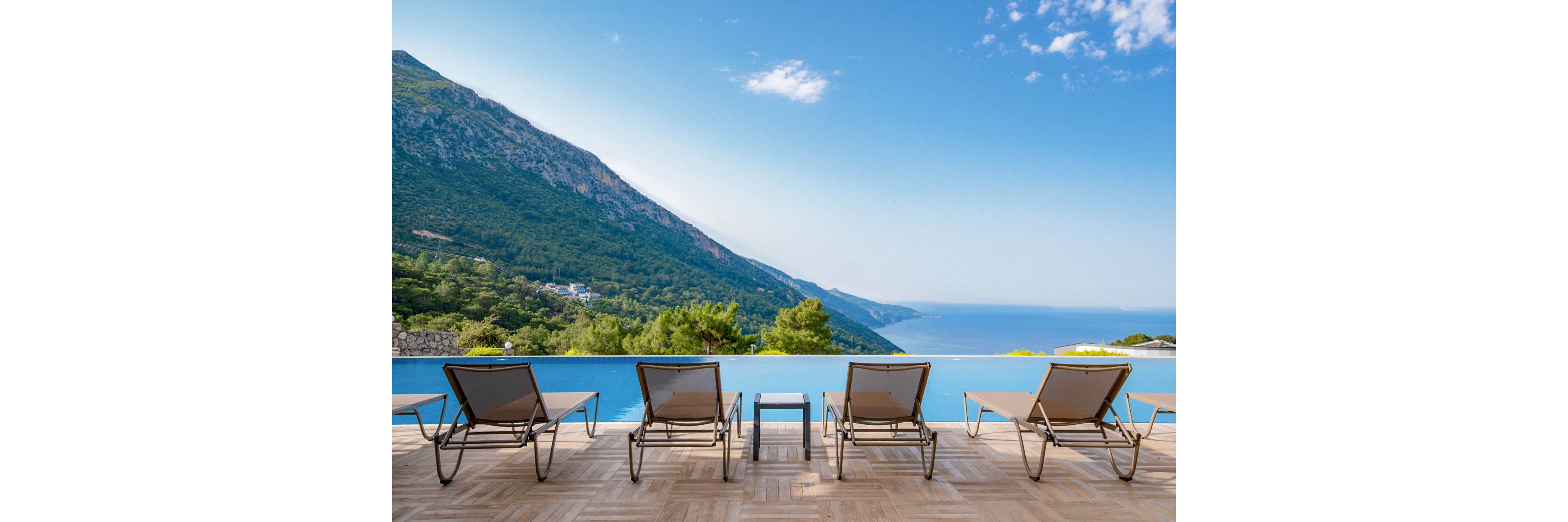 Four empty lounge chairs line the edge of an infinity pool facing a vast coastal landscape. A steep green mountain rises on the left, while the sea stretches into the distance under a clear blue sky. The pool seamlessly blends with the horizon, creating a tranquil and luxurious resort atmosphere. Warm wooden decking and minimal furniture emphasize relaxation and open space.