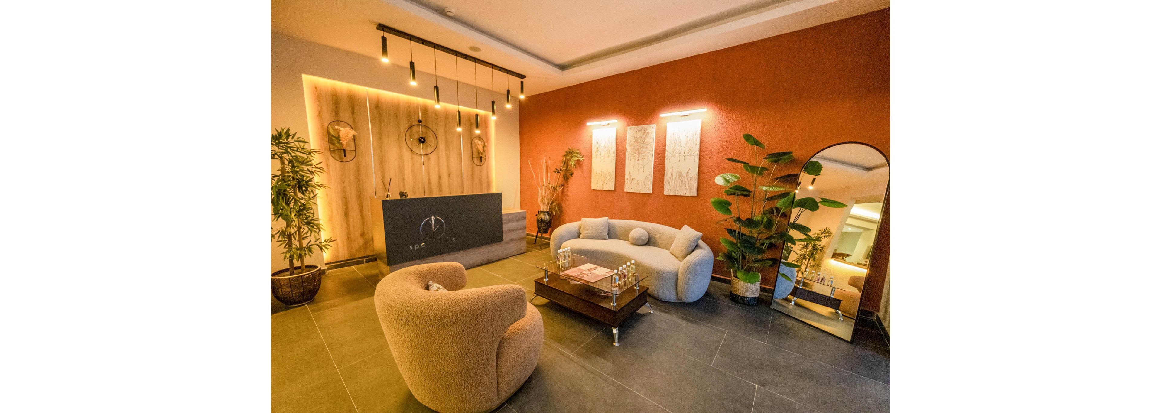 A contemporary lounge area features rounded sofas and armchairs arranged around a low wooden coffee table. Warm pendant lights hang above a minimalist reception counter set against a wooden accent wall. The space includes large potted plants, decorative wall art, and a tall floor mirror reflecting the room. Earthy tones and soft textures create a welcoming, relaxing atmosphere suitable for a lobby or waiting area.