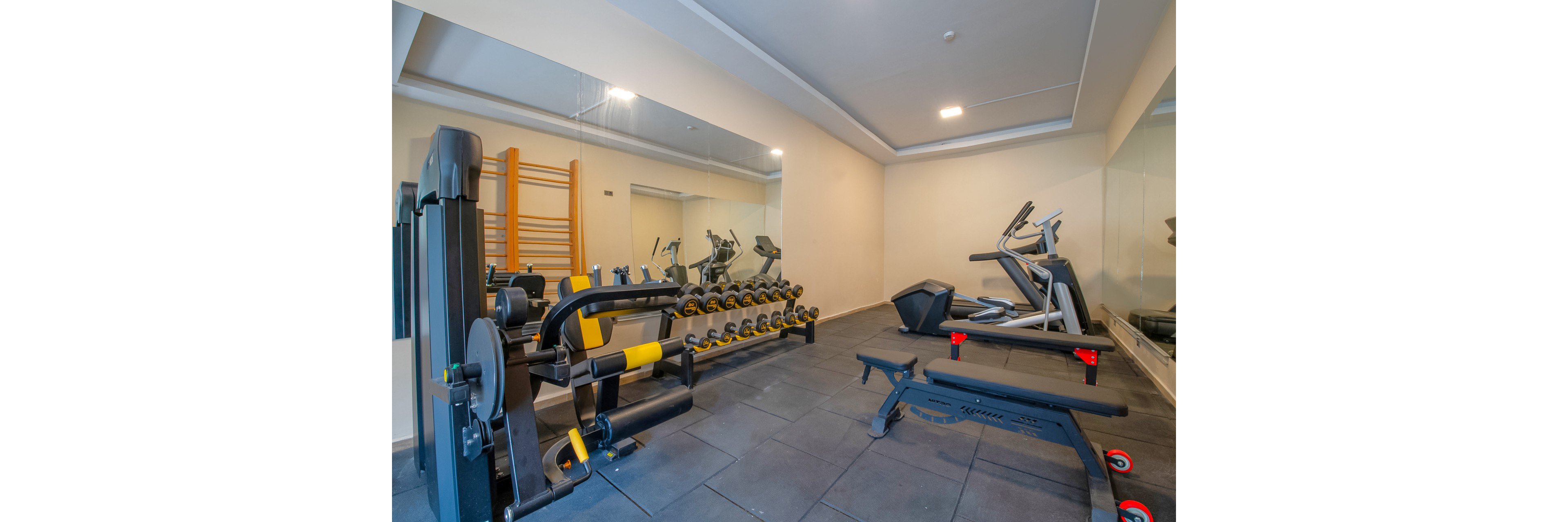 A compact modern gym room features various strength and cardio machines neatly arranged on a dark rubber floor. Free weights are organized on a rack in front of a large wall mirror, with a weight bench positioned nearby. In the background, treadmills and other cardio equipment face the mirrored wall, while a wooden wall ladder adds functional training options. Bright overhead lighting and neutral-colored walls create a clean, professional fitness environment.