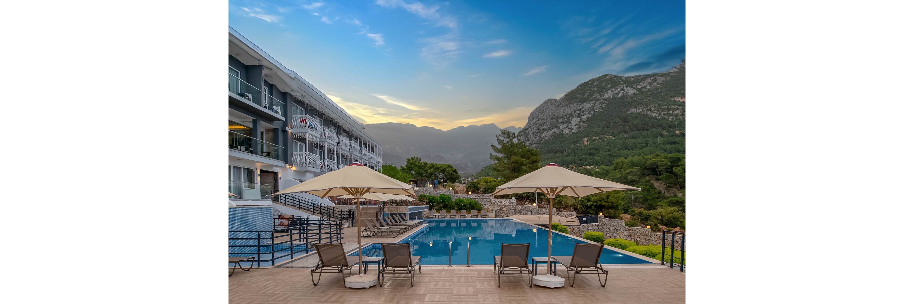 A modern resort swimming pool is set against a dramatic mountain landscape at dusk. Lounge chairs and large white umbrellas line the poolside deck, facing the calm blue water. A multi-story hotel building with balconies stands to the left, while lush green trees and rocky hills rise in the background. The sky shows soft evening light with scattered clouds, creating a tranquil vacation atmosphere.