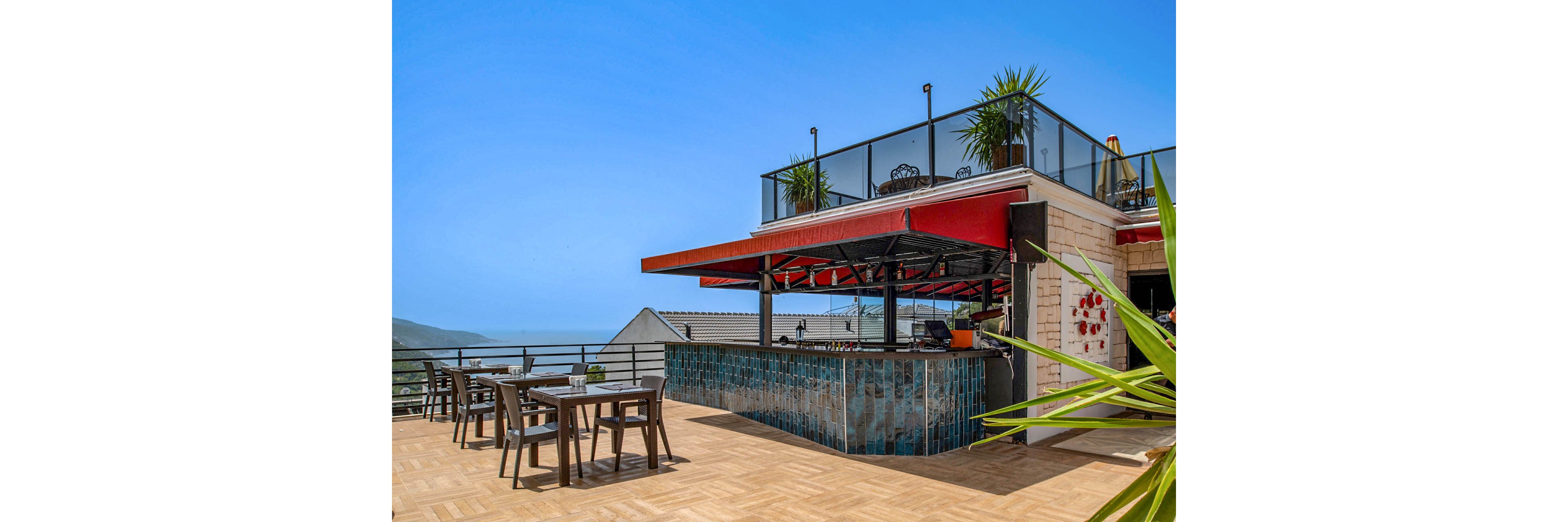 An open-air rooftop bar sits on a spacious terrace overlooking a distant coastline. The bar features blue-tiled fronts, a red awning, and modern glass railings around the upper level. Several empty tables and chairs are arranged on the wooden deck under a clear blue sky. Potted plants add greenery and a relaxed, contemporary atmosphere to the scene.