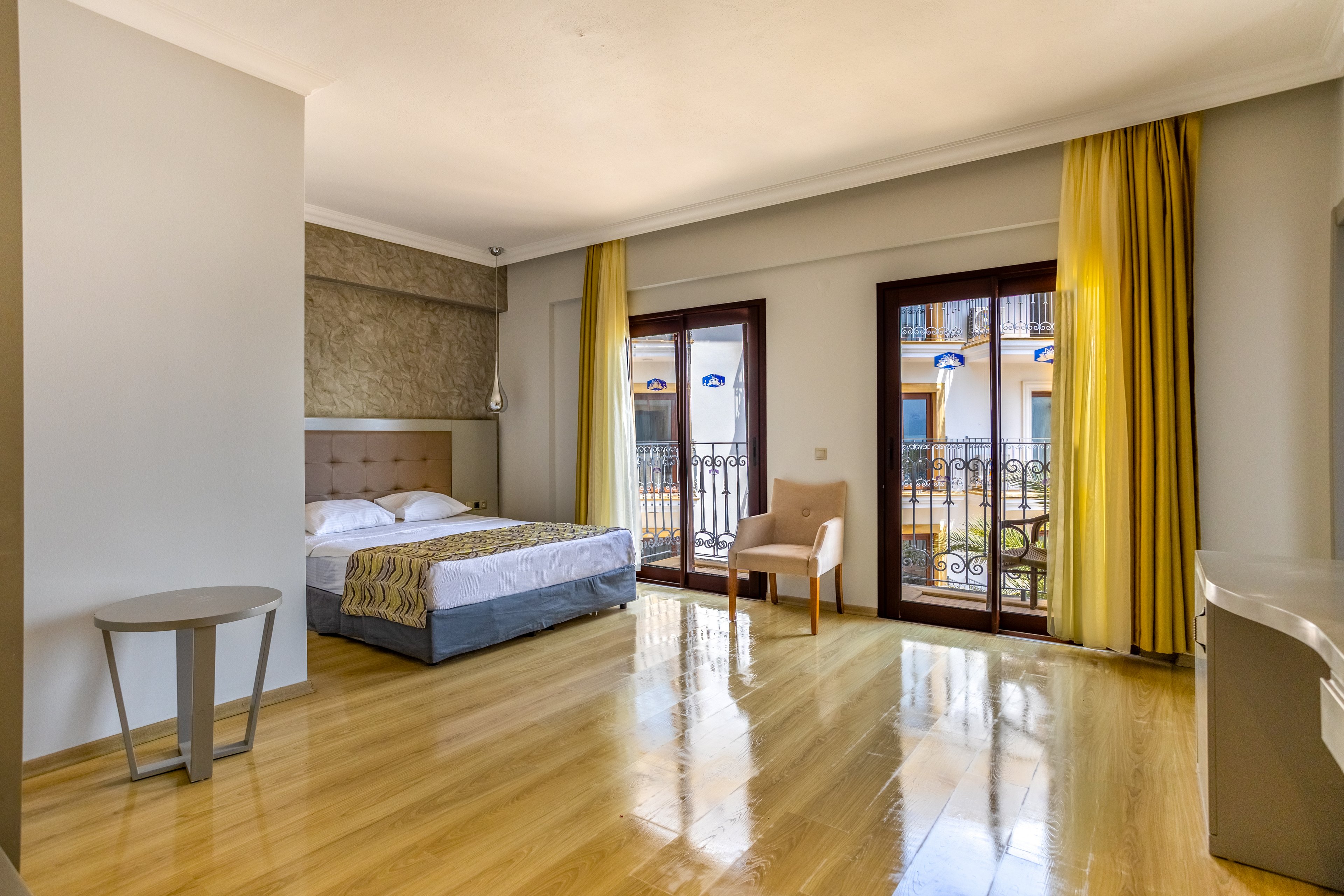 Spacious modern hotel room featuring a double bed with neutral bedding and a textured accent wall. Large glass doors open onto a balcony with decorative railings and views of neighboring buildings. Yellow curtains and light wooden flooring create a warm, inviting atmosphere. A simple chair, side table, and desk complete the minimalist interior design.