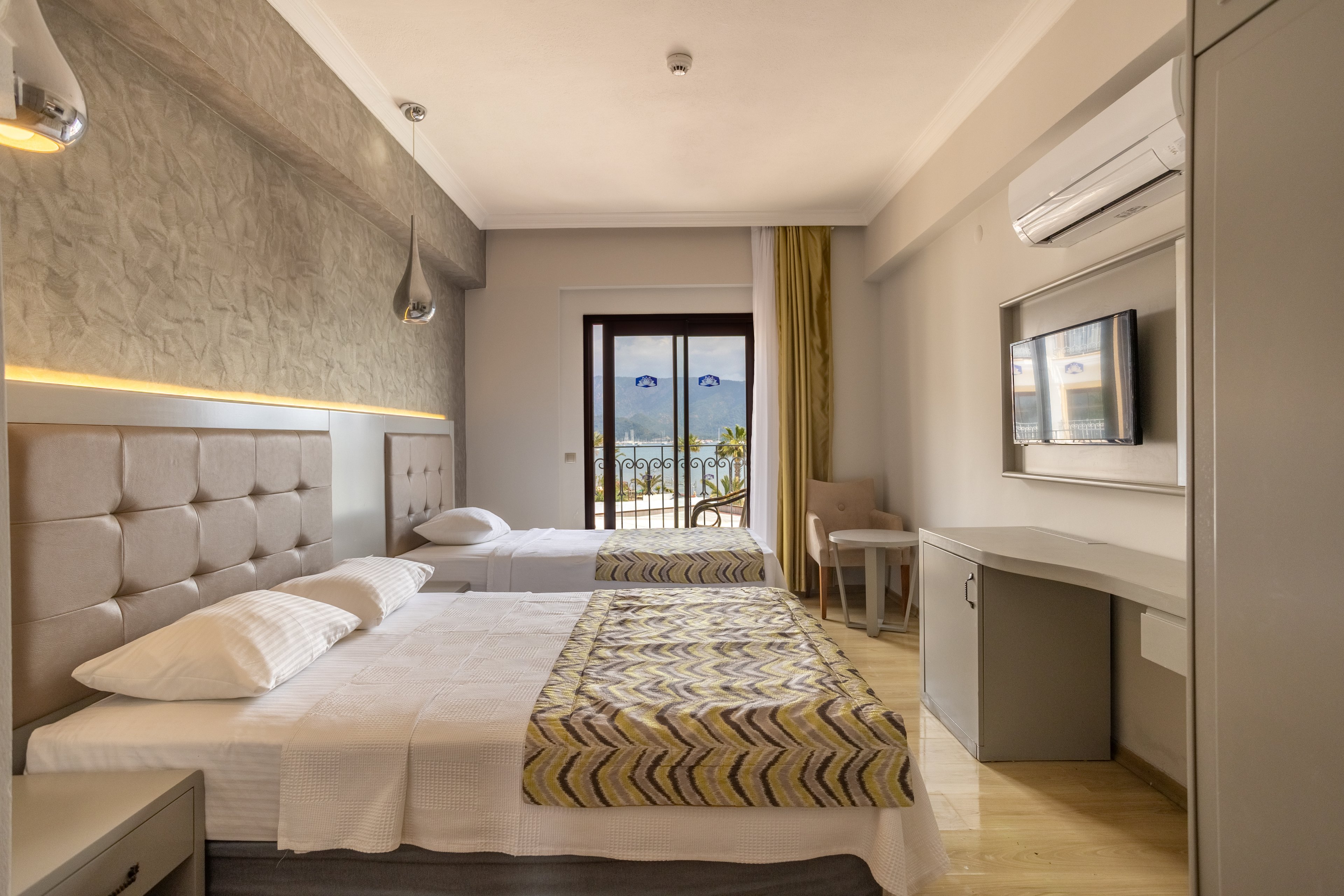 A contemporary hotel room features a double bed and a single bed with patterned bed runners. The space includes a wall-mounted television, desk, chair, and air conditioning unit for guest comfort. Warm neutral tones, soft lighting, and minimalist decor create a calm, inviting atmosphere. A glass door opens to a balcony with a scenic outdoor view of mountains and water in the distance.