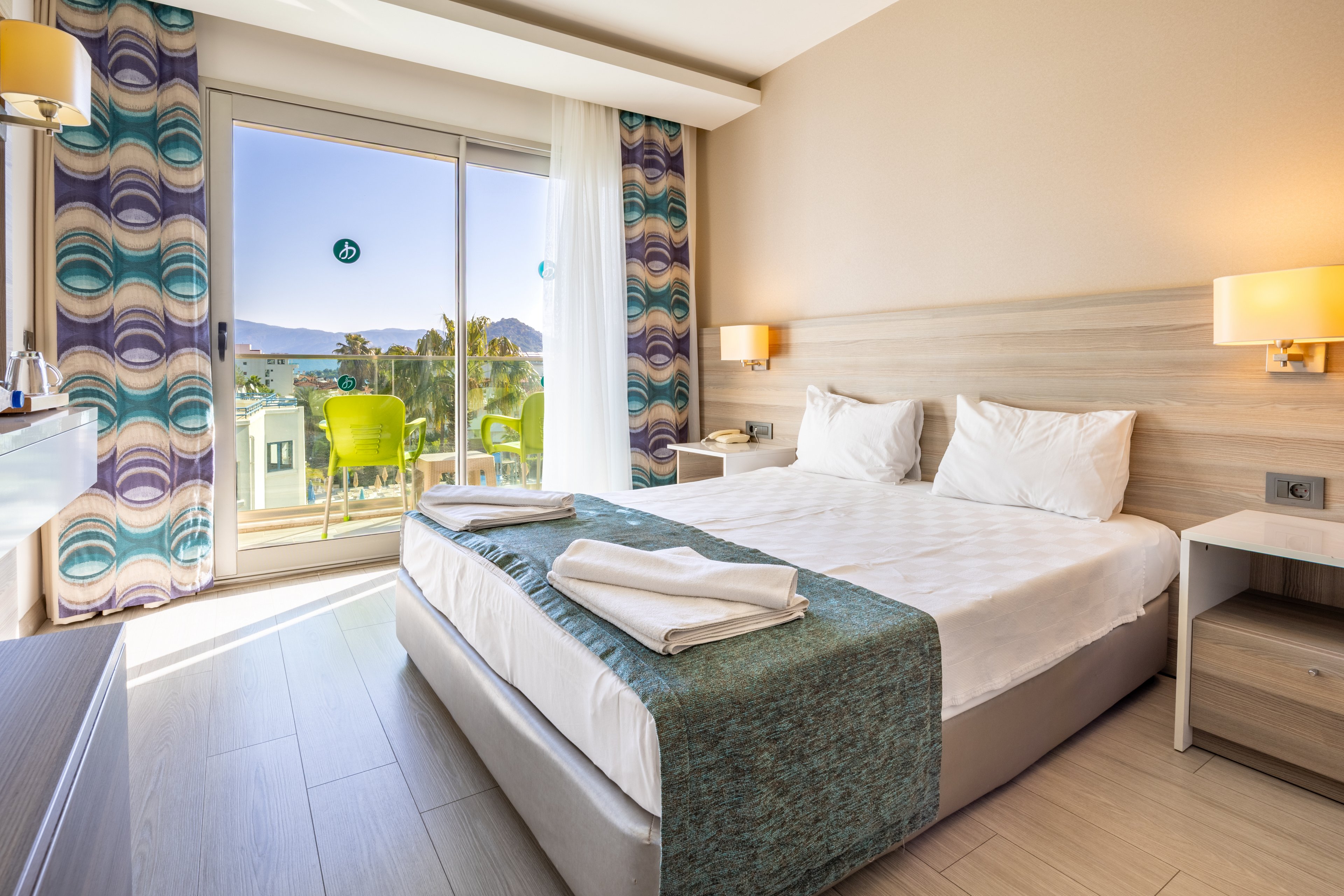 A modern hotel room features a neatly made double bed with white linens and teal accents. The space is filled with natural light from large sliding glass doors that open to a balcony overlooking the sea and distant mountains. The room includes contemporary decor, patterned curtains, and soft wall lighting, creating a fresh and inviting atmosphere. Two towels are neatly folded on the bed, and the overall palette is light and airy.